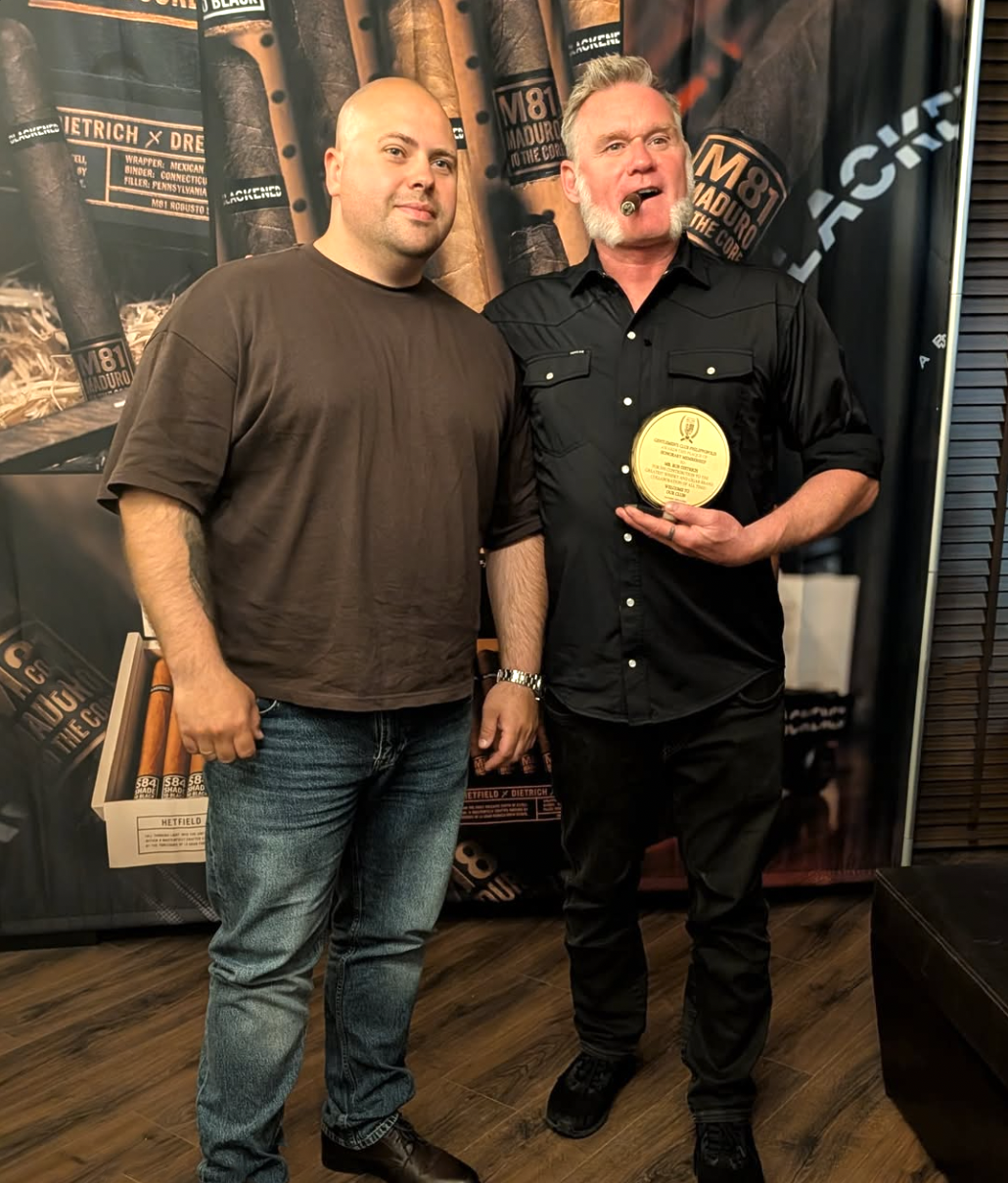 Two men standing indoors, one holding a round award or medal, the other smoking a cigar, with a backdrop featuring cigars.