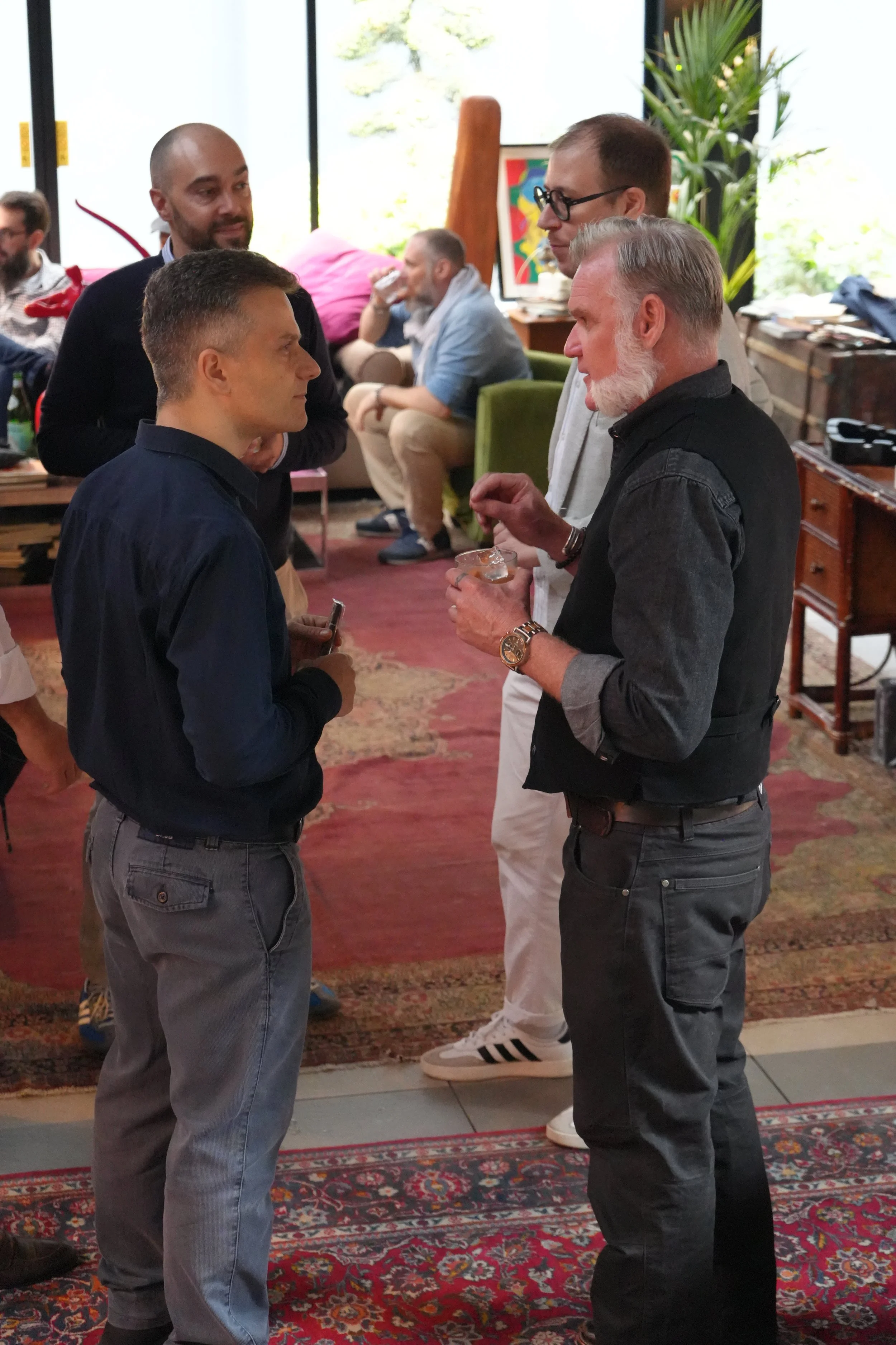 Four men engaged in conversation at a social gathering, with artwork on a TV screen in the background and a patterned rug on the floor.
