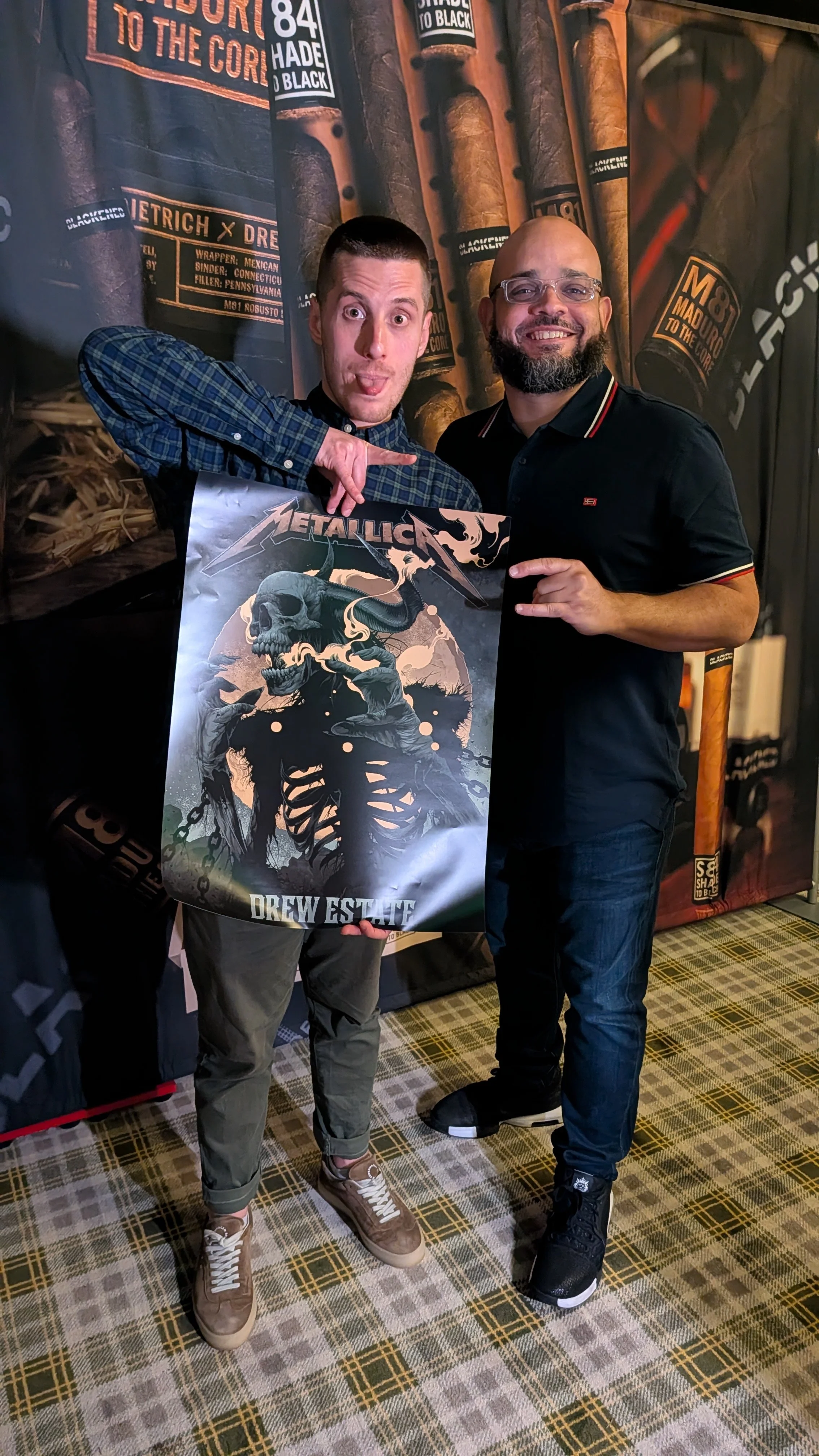 Two men standing indoors, one holding a 'Metallica Drew Estate' poster, smiling at the camera. Background has cigar-themed banners and a carpeted floor with a checkered pattern.