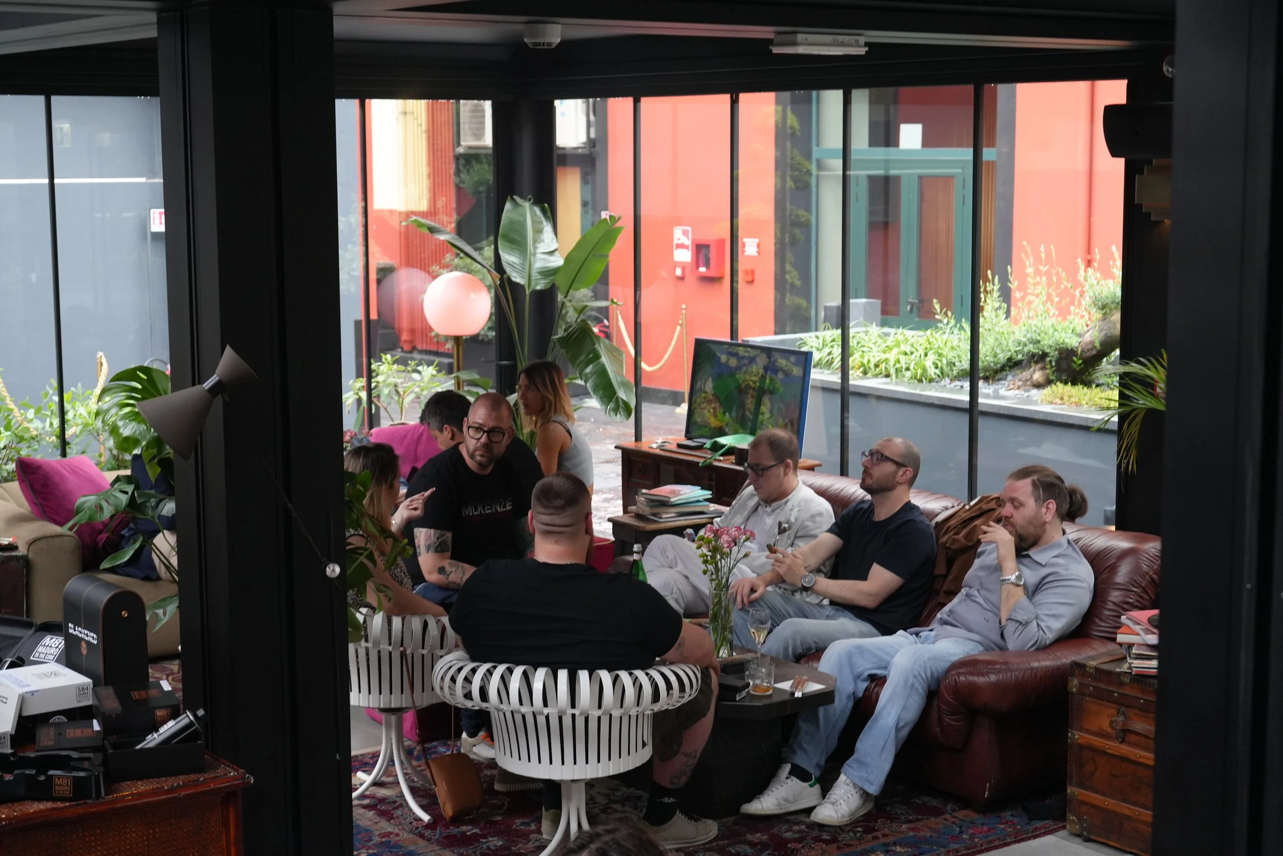 A group of people sitting in a lounge or café with large glass windows showing an outdoor garden and red building, decorated with plants, a globe lamp, and books.
