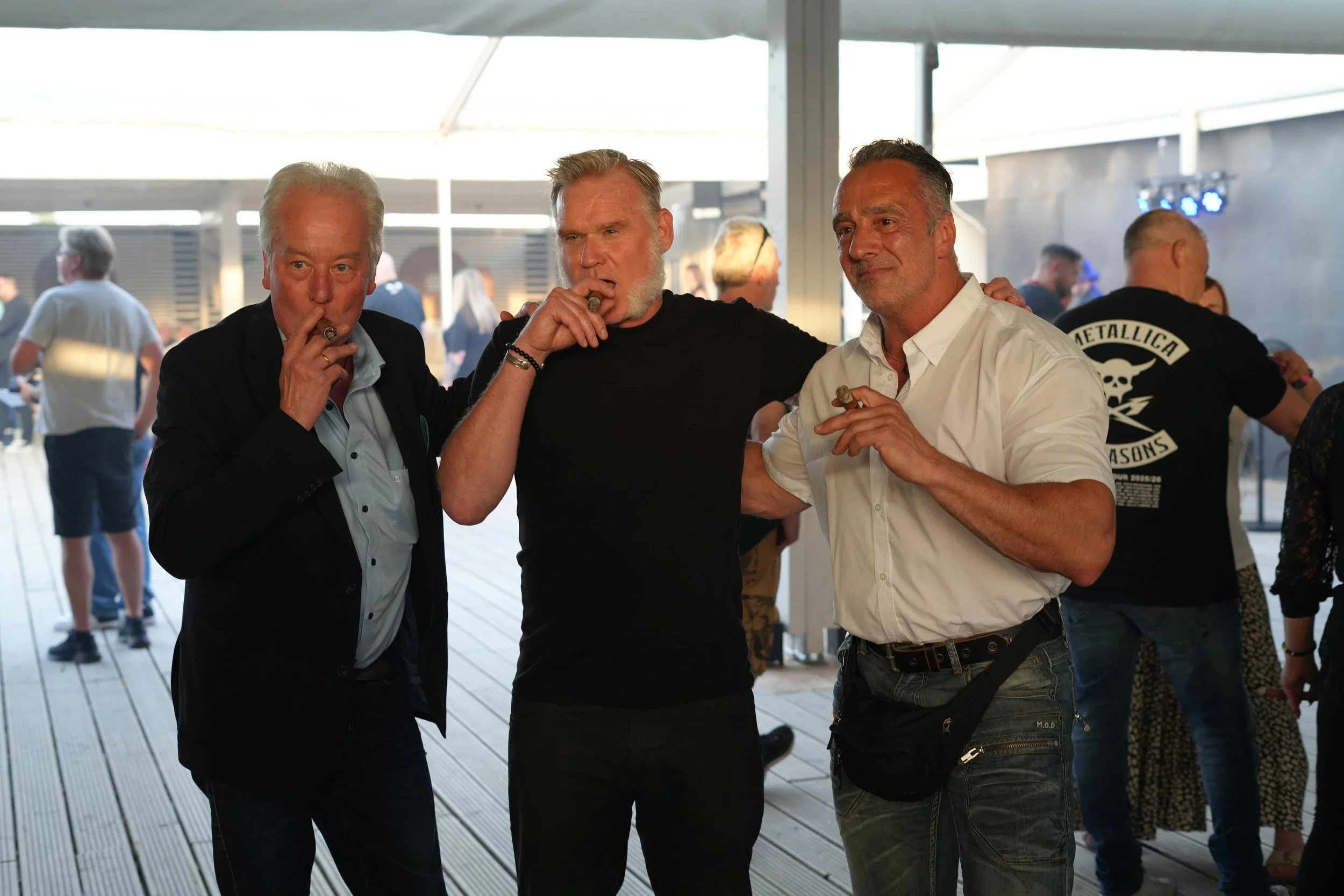 Three men standing together at a social event, smoking cigars, with a background of other people in a well-lit indoor or semi-outdoor setting.