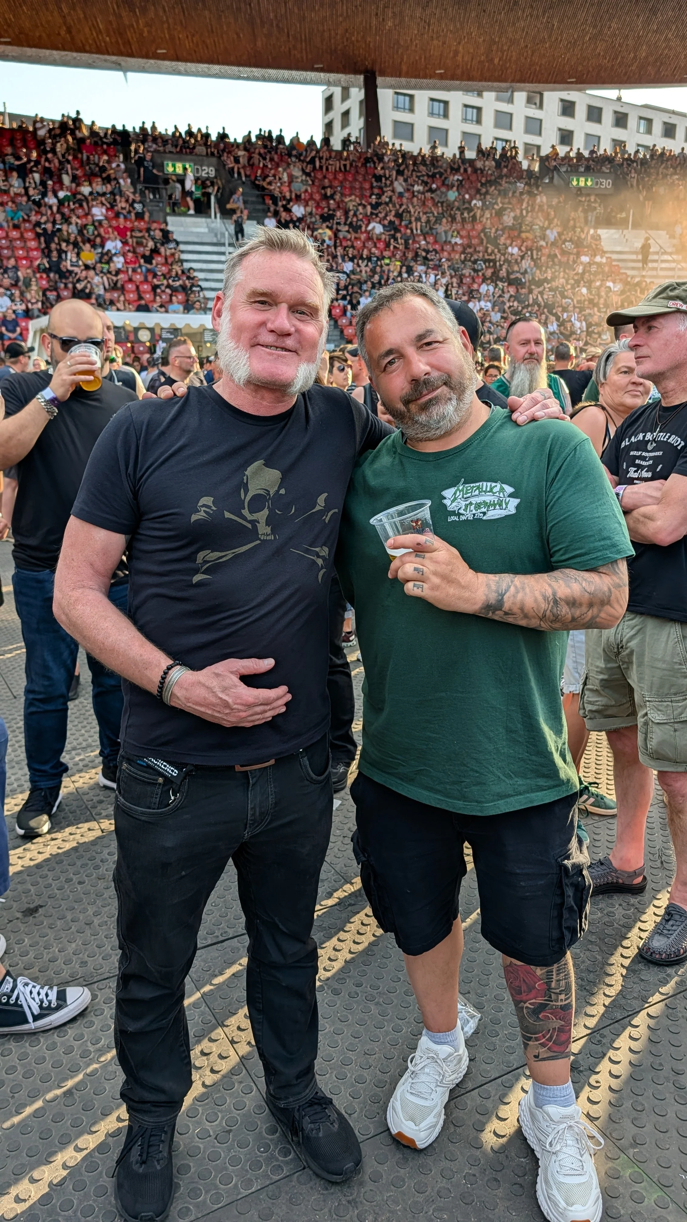 Two men posing together at a Metallica concert. One man is wearing a black T-shirt with a skull graphic, and the other is wearing a green Metallica T-shirt and holding a plastic cup. Both men are smiling and have their arms around each other.