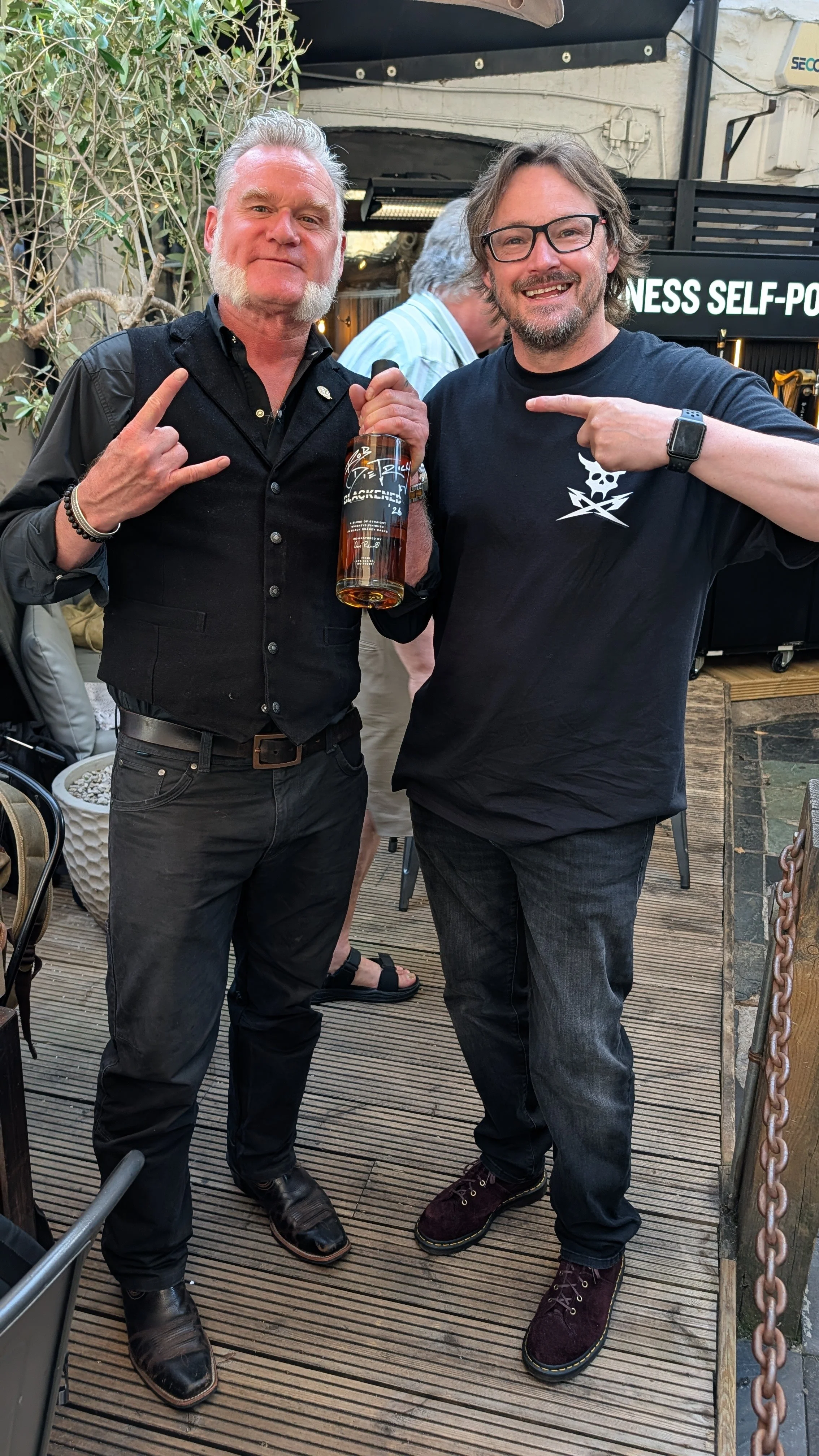 Two men standing on a wooden deck at an outdoor event, smiling and pointing at each other, one holding a whiskey bottle, with a black sign that says 'BUSINESS SELF-PO' partially visible in the background.