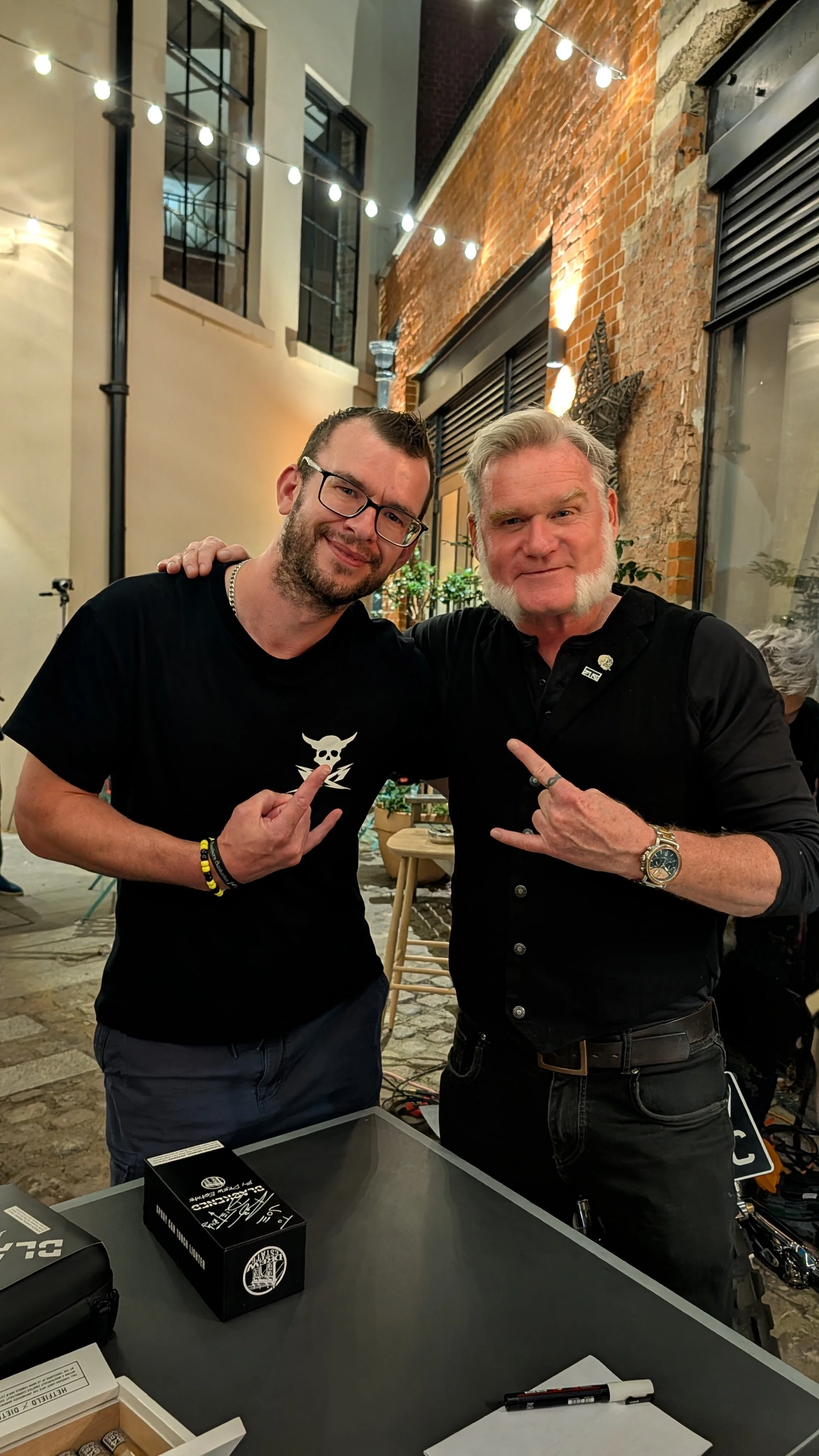 Two men at a night outdoor event, making hand signs and smiling, with string lights and brick walls in the background.