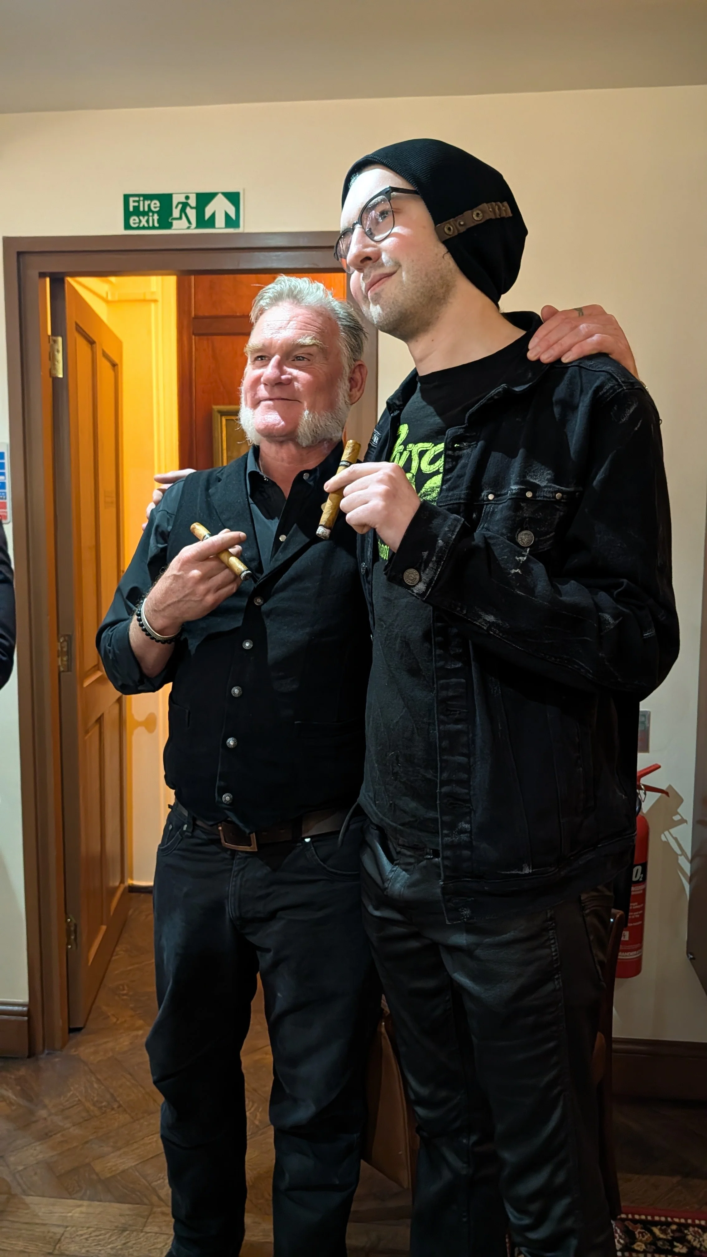 Two men stand indoors, one with a beard and gray hair, the other younger with glasses and a beanie, both holding cigars and smiling. There is a fire exit sign above a door in the background.