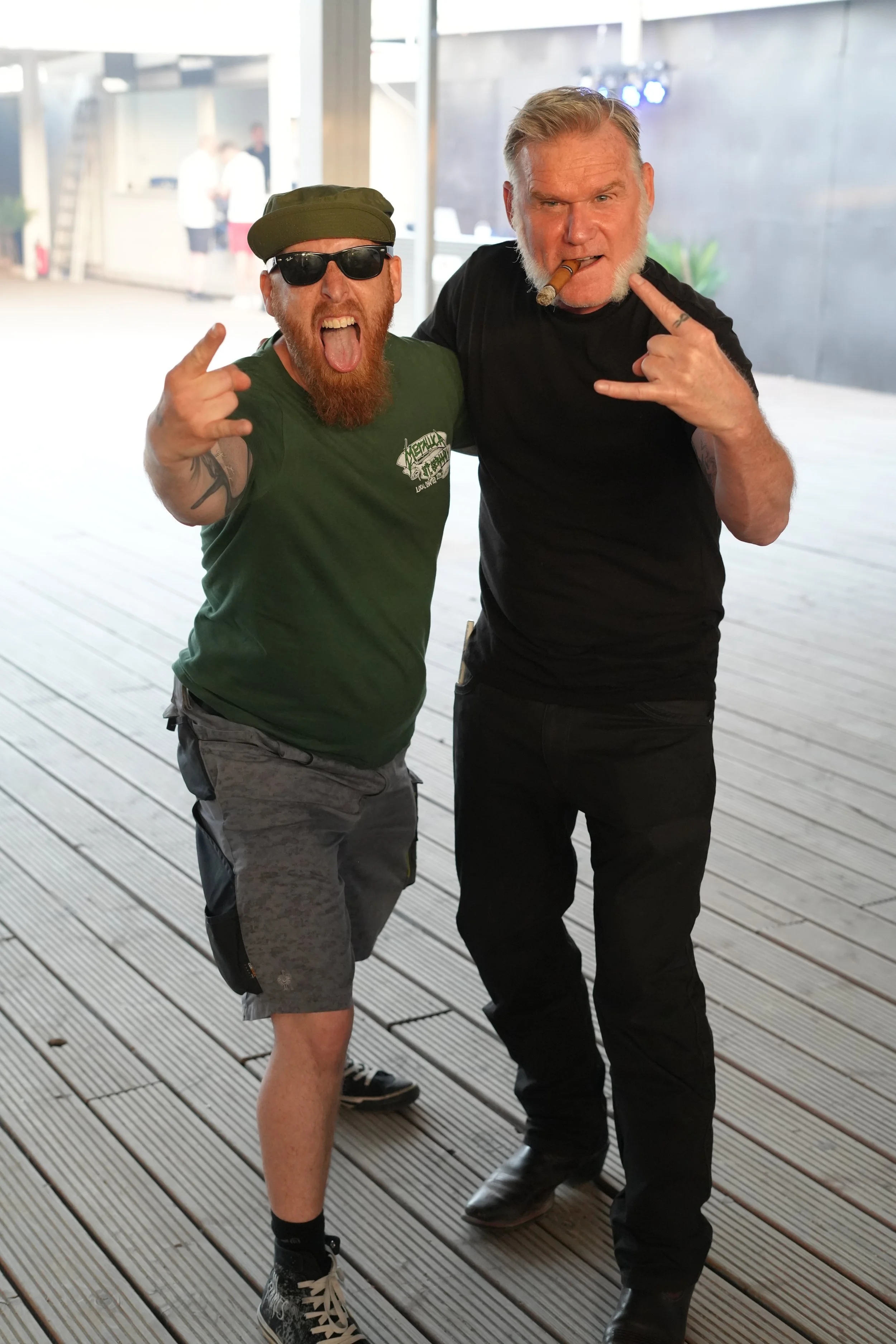 Two men posing with rebellious gestures and expressions on a wooden deck indoors. One man wears sunglasses, a green cap, and a green t-shirt. The other man has a cigar in his mouth, a black t-shirt, and makes a rock 'n' roll hand sign.