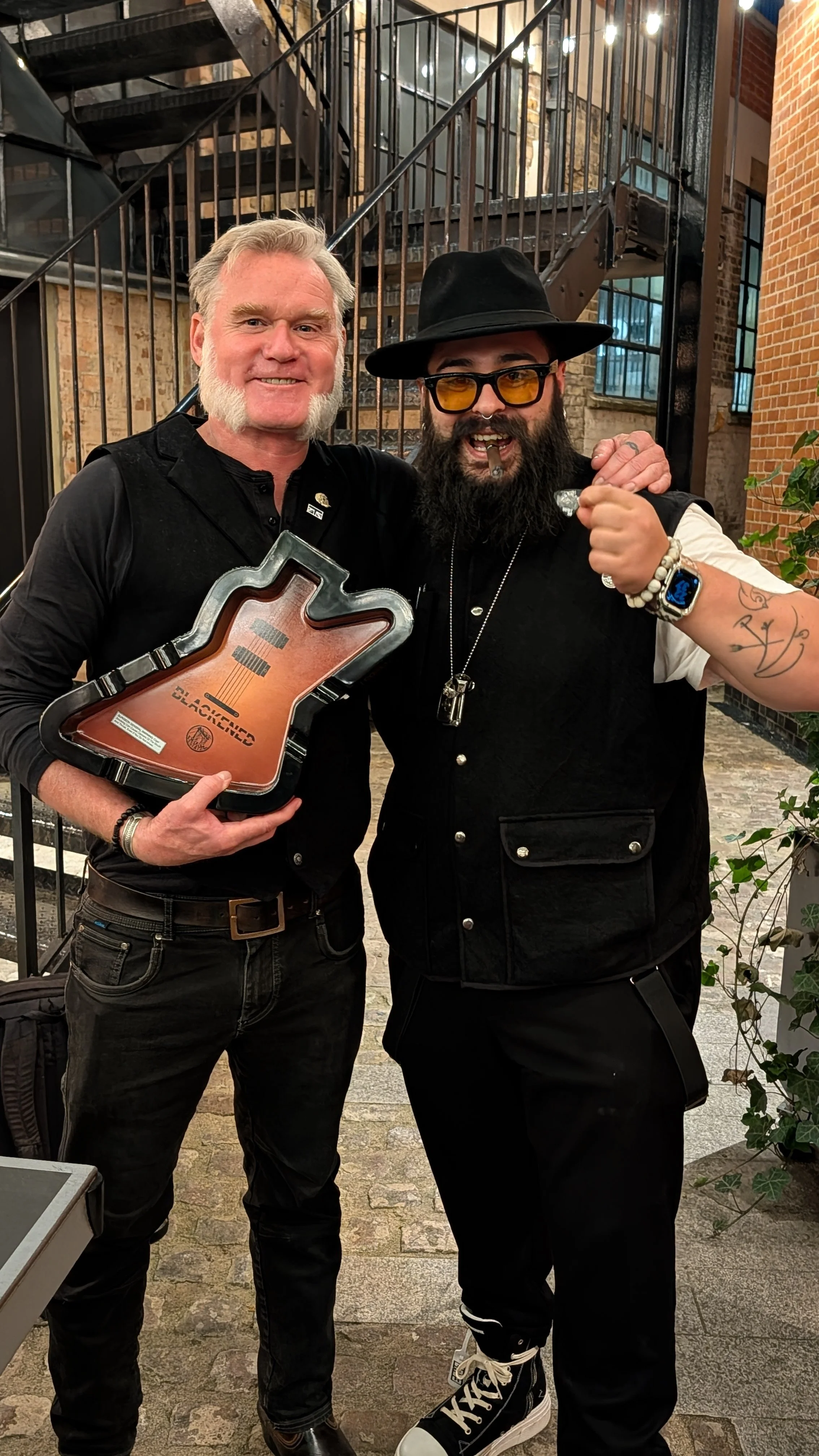 Two men standing together in an industrial-style location with exposed brick and a metal staircase. The man on the left has gray hair and a beard, wearing black clothing, and holding a guitar-shaped award. The man on the right has a full beard, weari