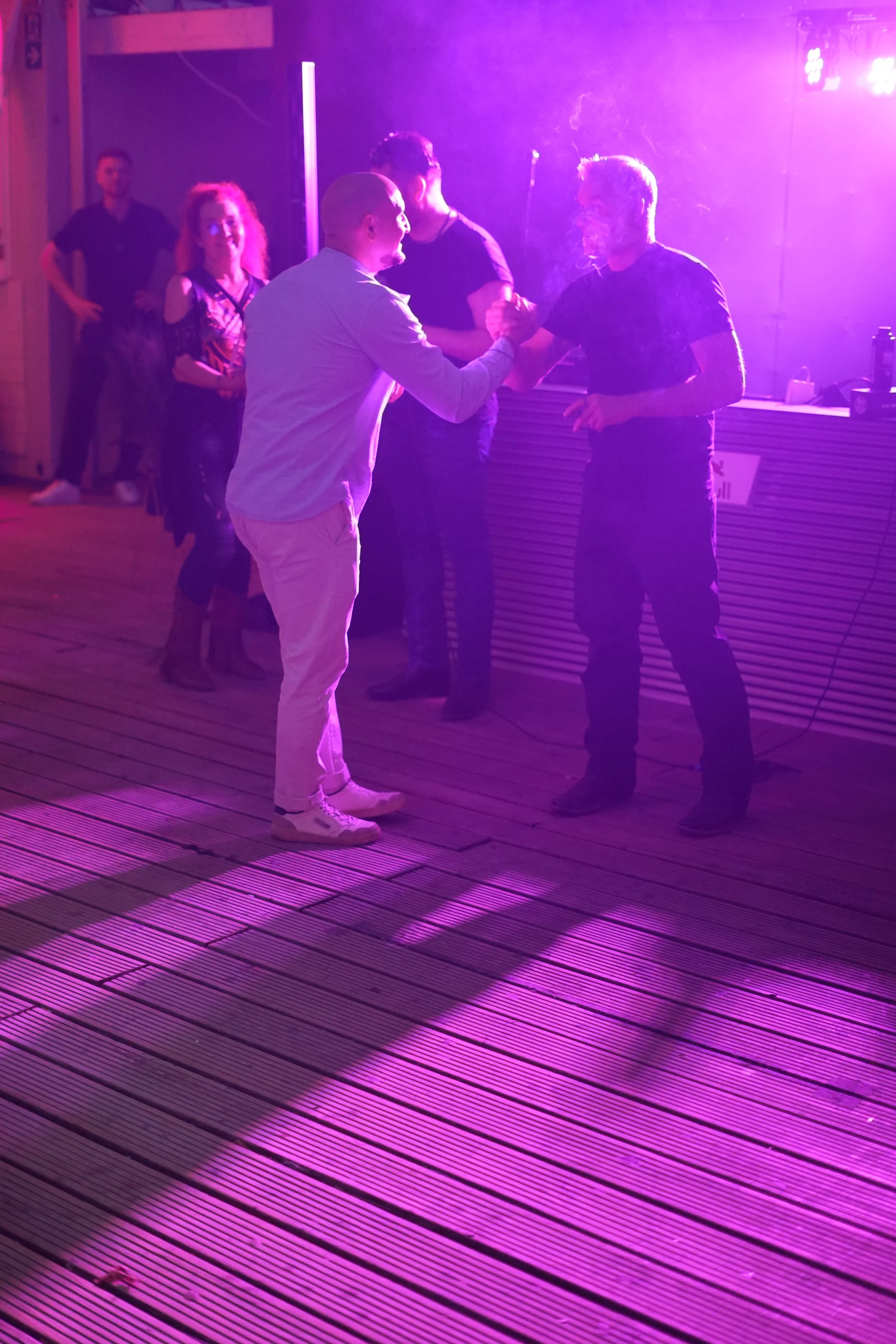 Two men shaking hands at a party or nightclub with purple lighting, with three women in the background against a wall.