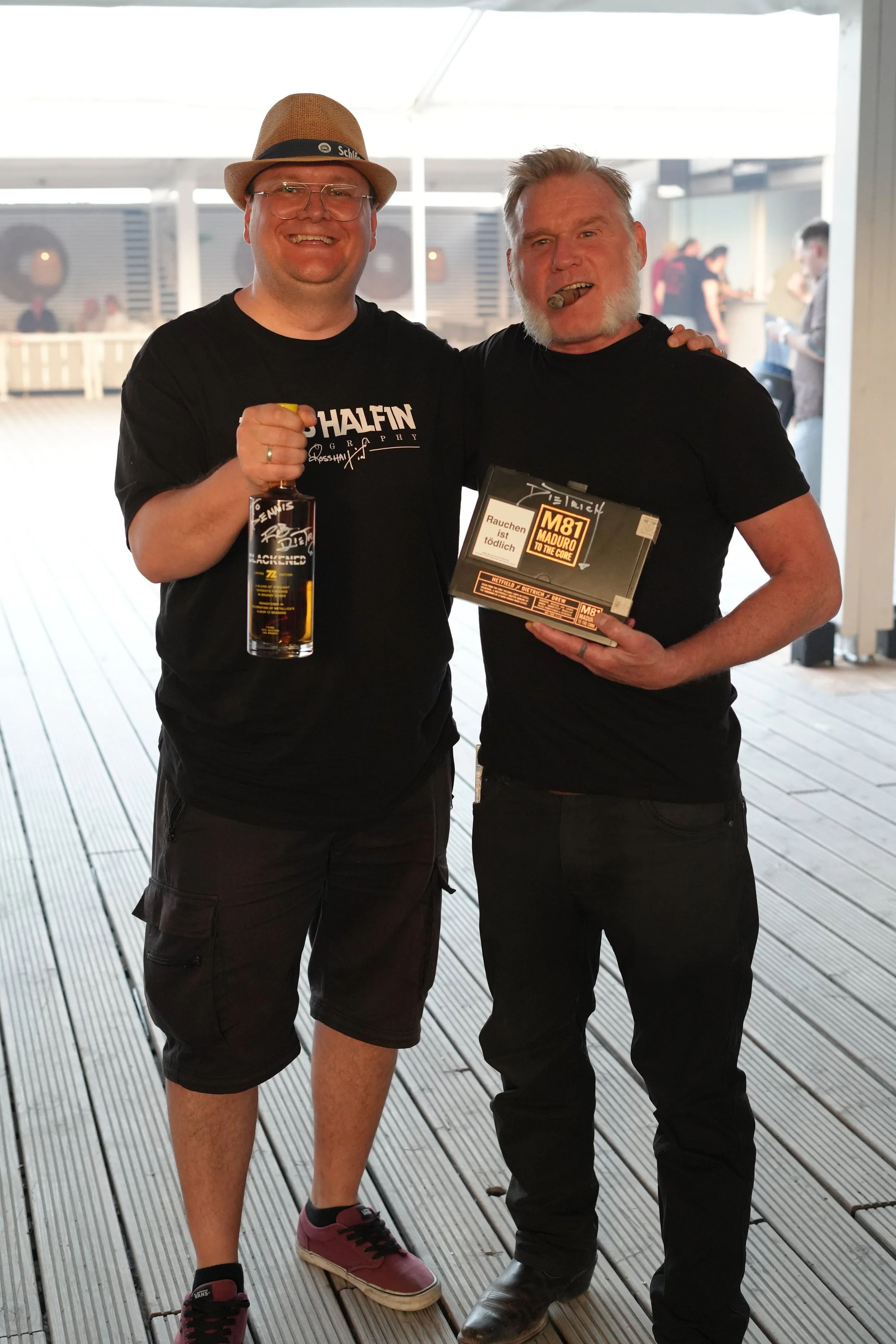 Two smiling men standing arm in arm inside a well-lit, semi-outdoor space with a wooden deck floor. The man on the left is wearing a tan fedora, glasses, a black T-shirt, black shorts, and pinkish sneakers, holding a bottle of alcohol. The man on the