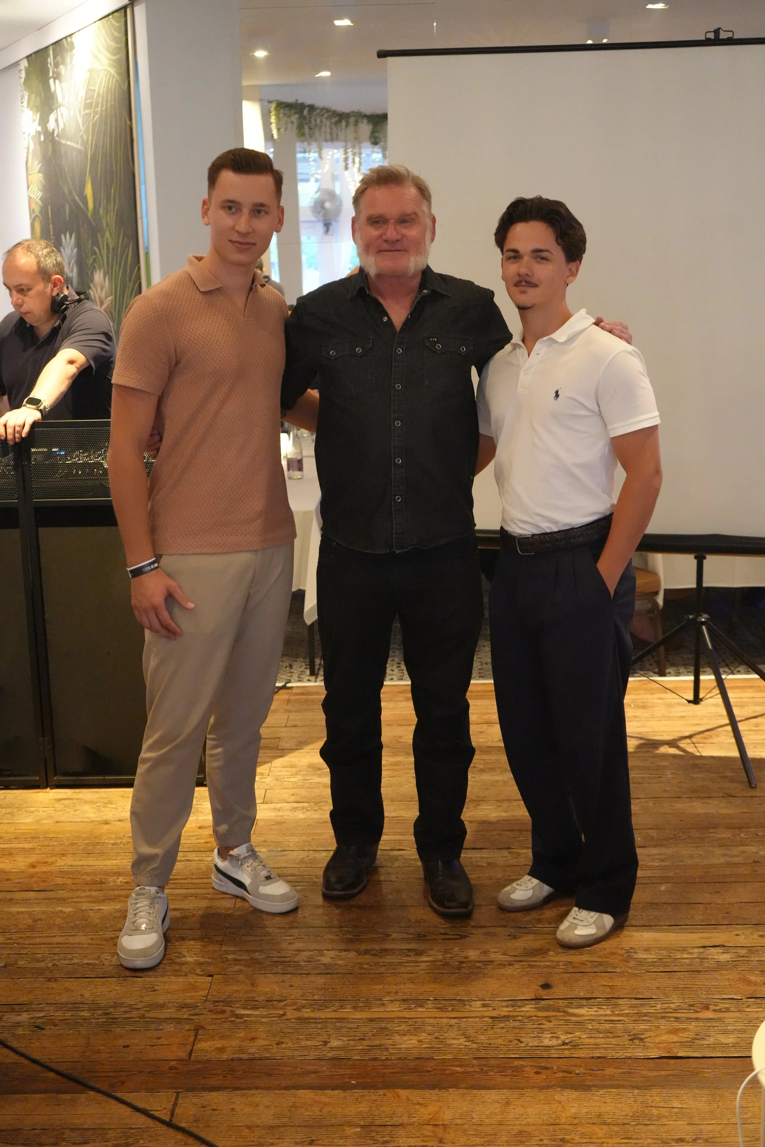 Three men standing side by side indoors with a DJ in the background. The men are smiling and posing for the photo.