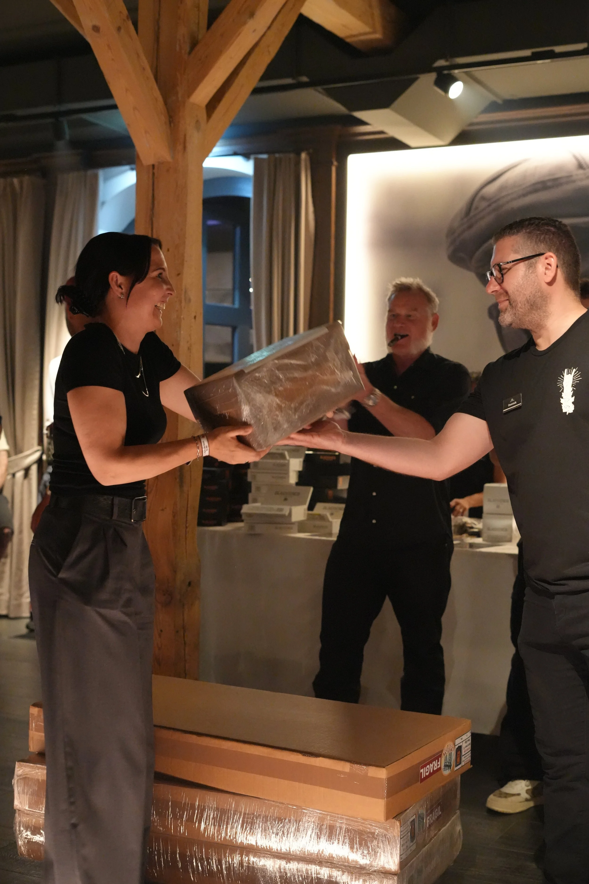 A woman is receiving a wrapped gift from a man at a gathering or celebration, with another man smiling in the background. There are boxes and a table with items behind them.
