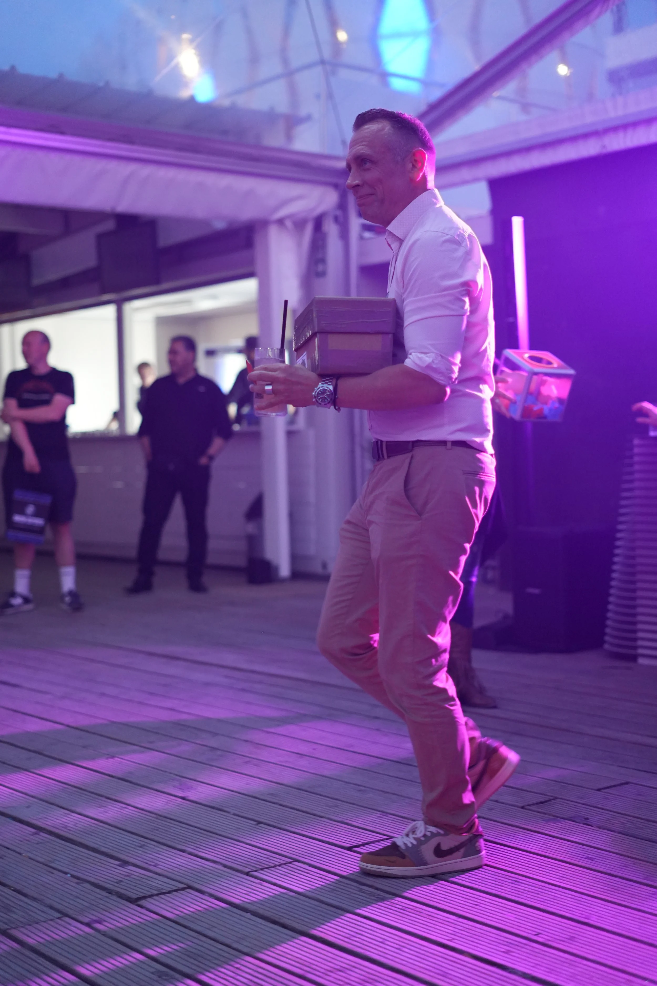 A man in a white shirt and beige pants holding a drink, a box, and another object, walking on a wooden floor illuminated with purple and blue lights during an indoor event.