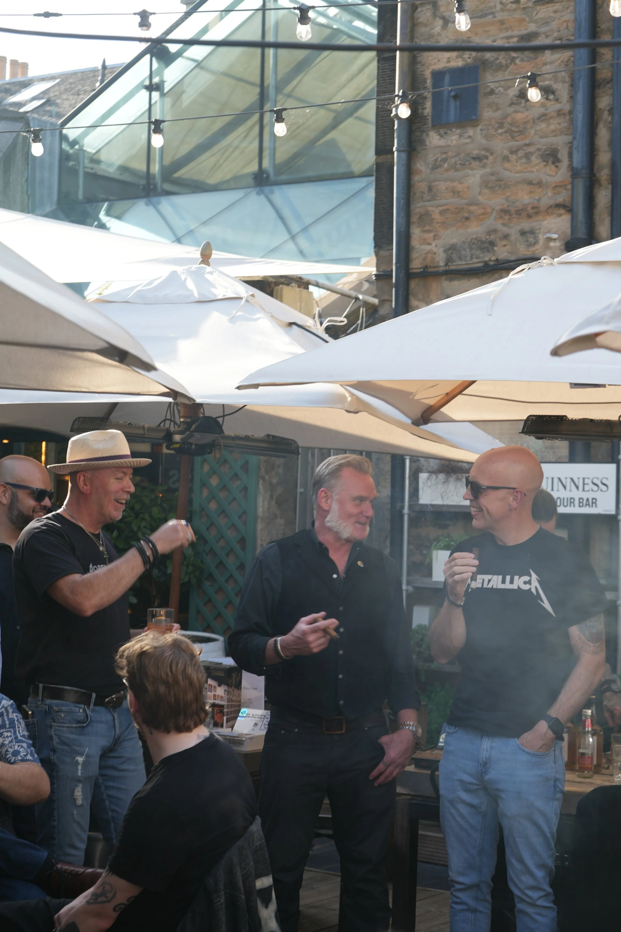 Group of men socializing outdoors under umbrellas with string lights overhead on a patio, with a brick building and glass structure in the background.