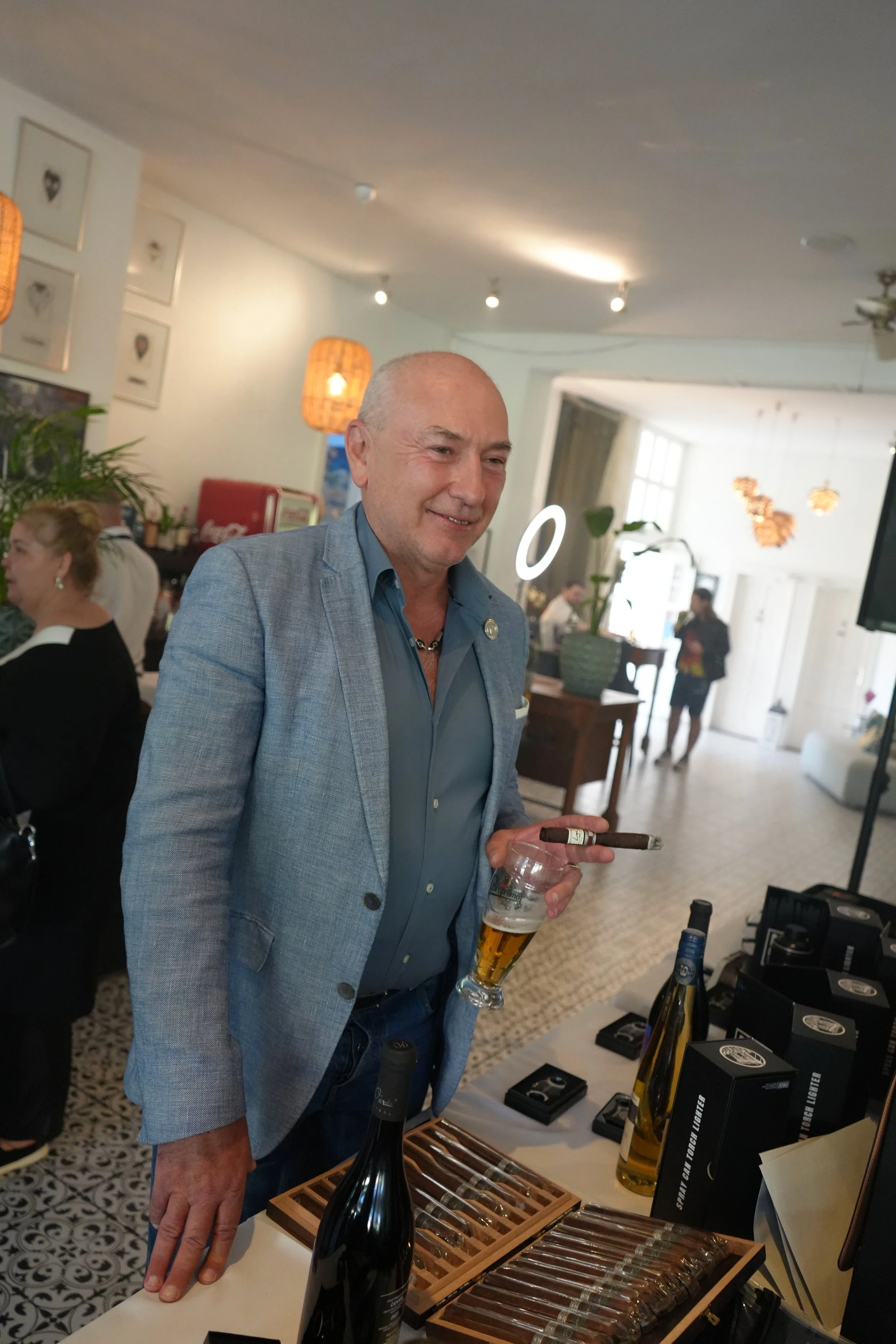 A man in a light blue blazer holding a cigar and a glass of beer stands indoors at a social gathering. The room is decorated with framed artwork, plants, and hanging lamps. There are people in the background, and a table in front of him displays bott