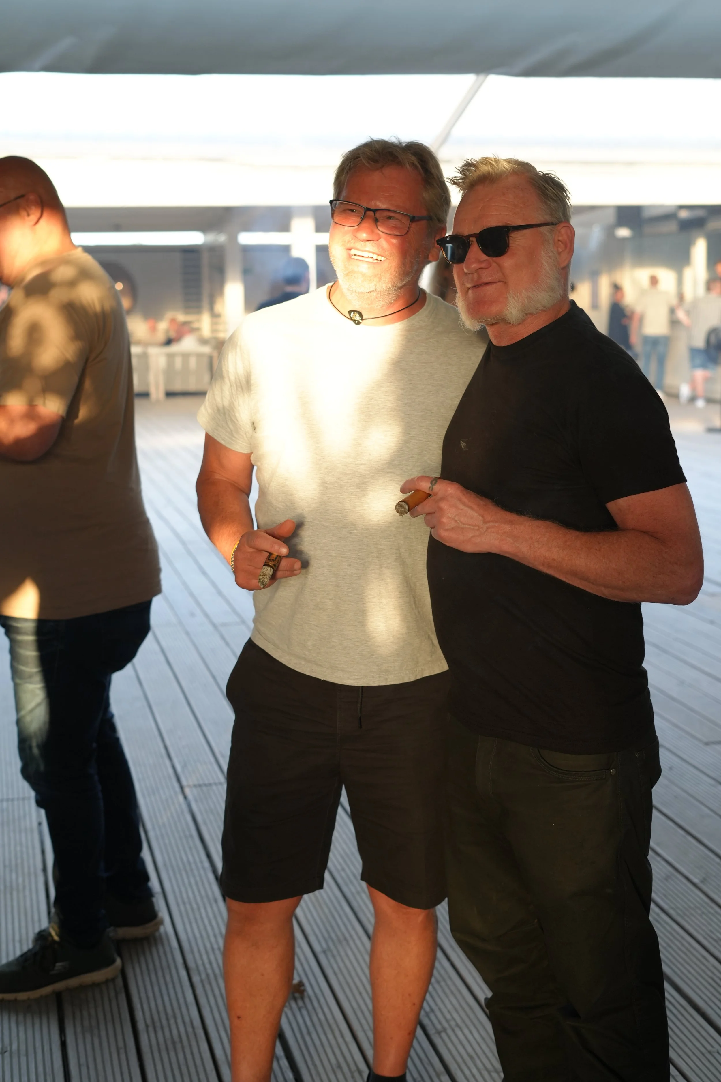 Two men standing outdoors on a boardwalk holding cigars, smiling, with other people in the background.