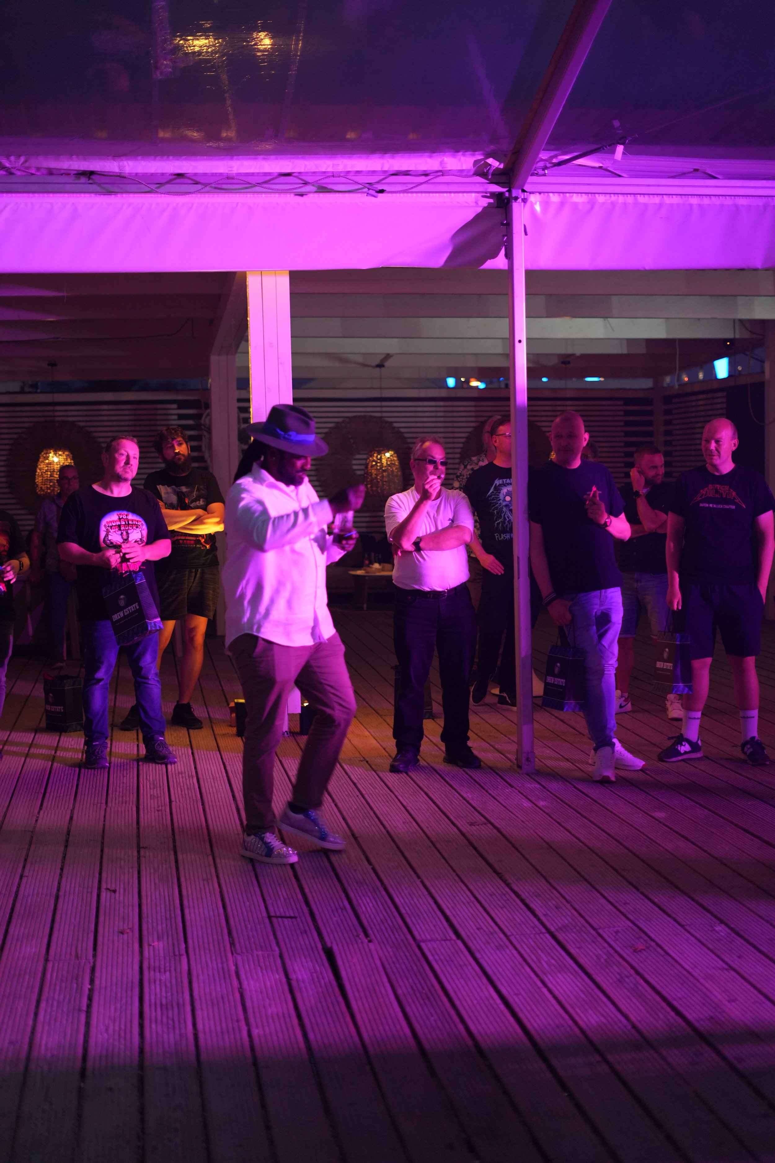 A group of people standing under purple and pink lighting on a wooden deck, some wearing casual clothing and sunglasses, with a vendor in the foreground wearing a hat, white shirt, and sneakers.