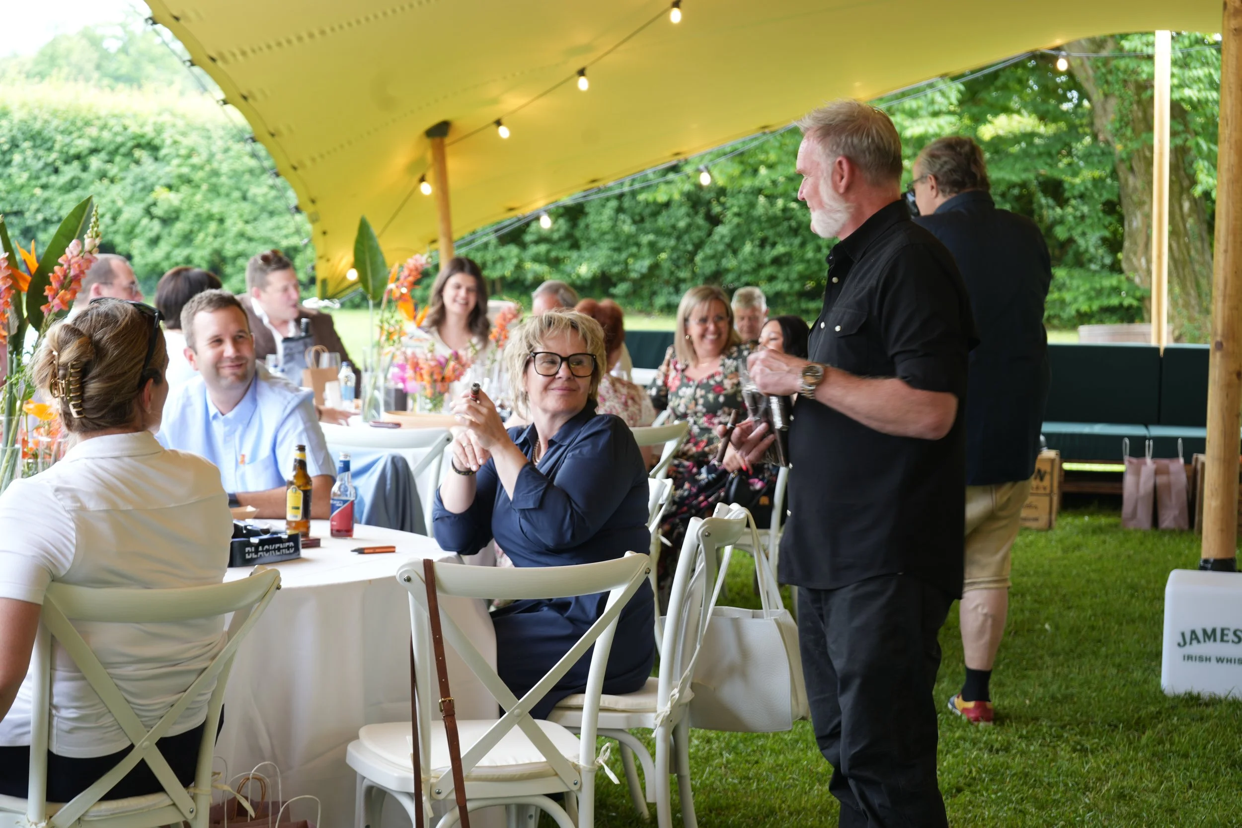 People gathered outdoors under a yellow tent, sitting at decorated tables, engaged in conversation, with a man standing and speaking to the group. Green trees are visible in the background.