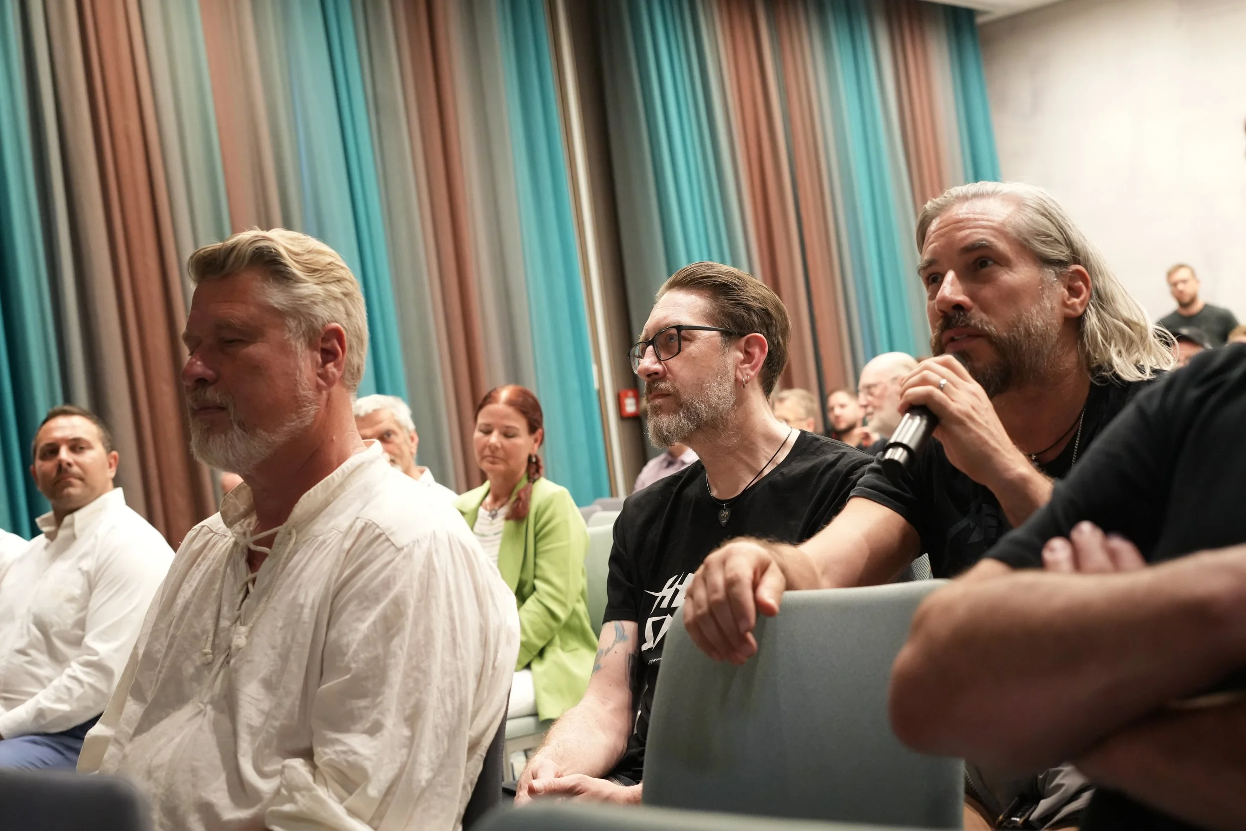 People attending a conference in a room with colorful curtains, some are seated and listening, one person with a beard and long hair is holding a microphone.