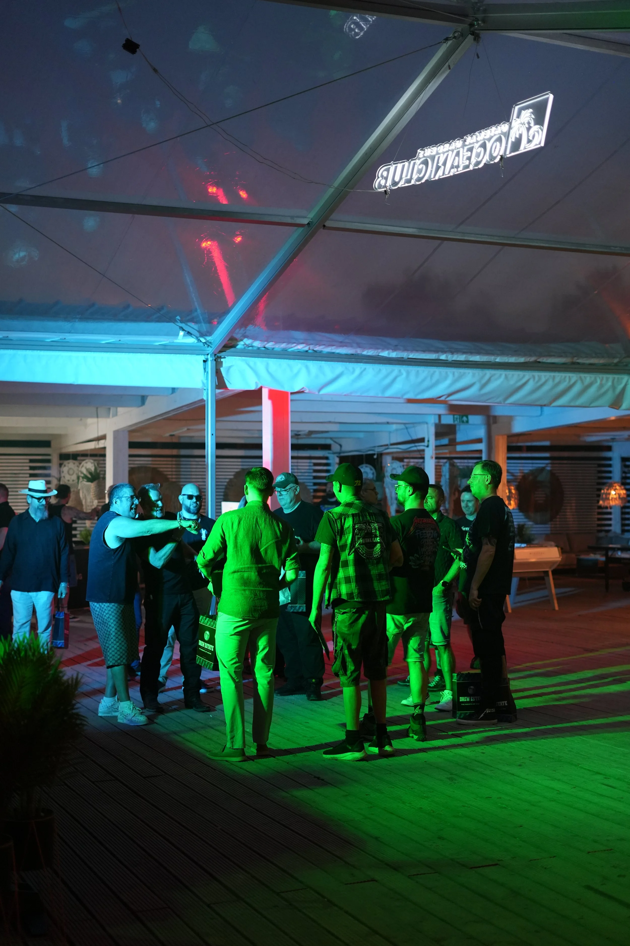 A group of people talking under a large outdoor canopy at night, with colorful lighting and a neon sign visible in the background.