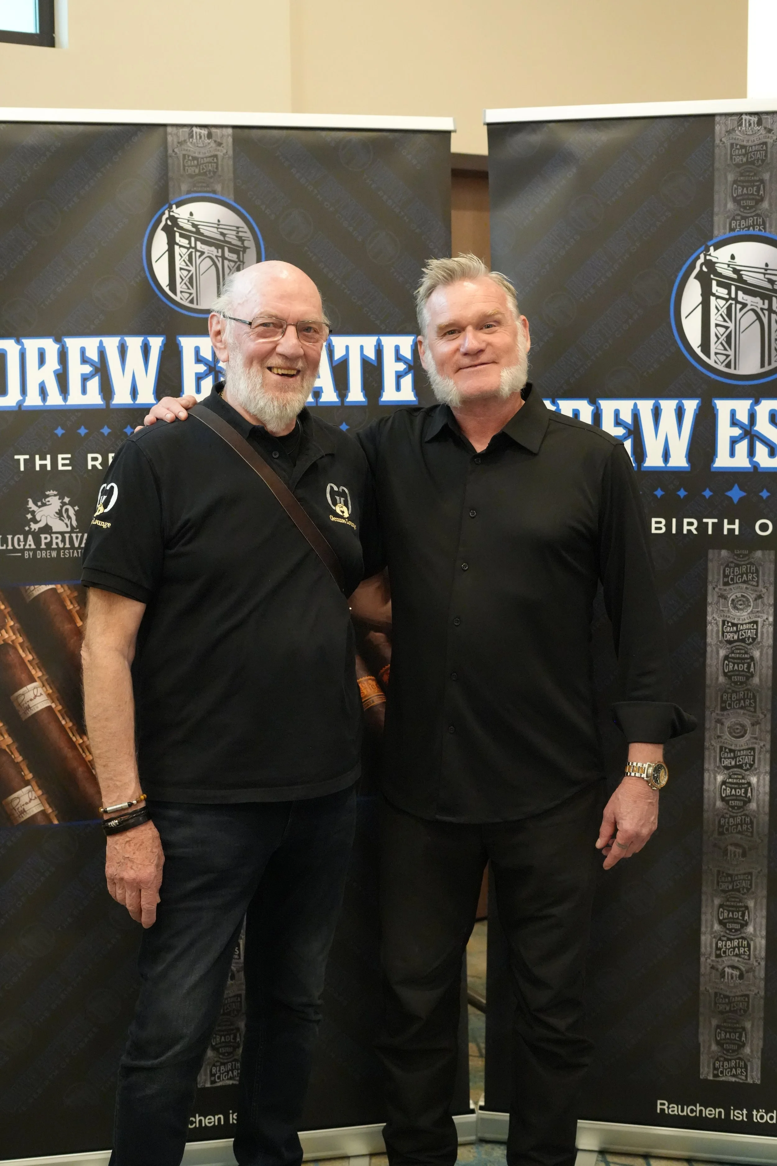 Two men standing in front of a Drew Estate banner, smiling at the camera. One man has glasses, a beard, and is wearing a black polo shirt with a logo; the other has styled hair and a goatee, wearing a black button-up shirt. Both are posing with their