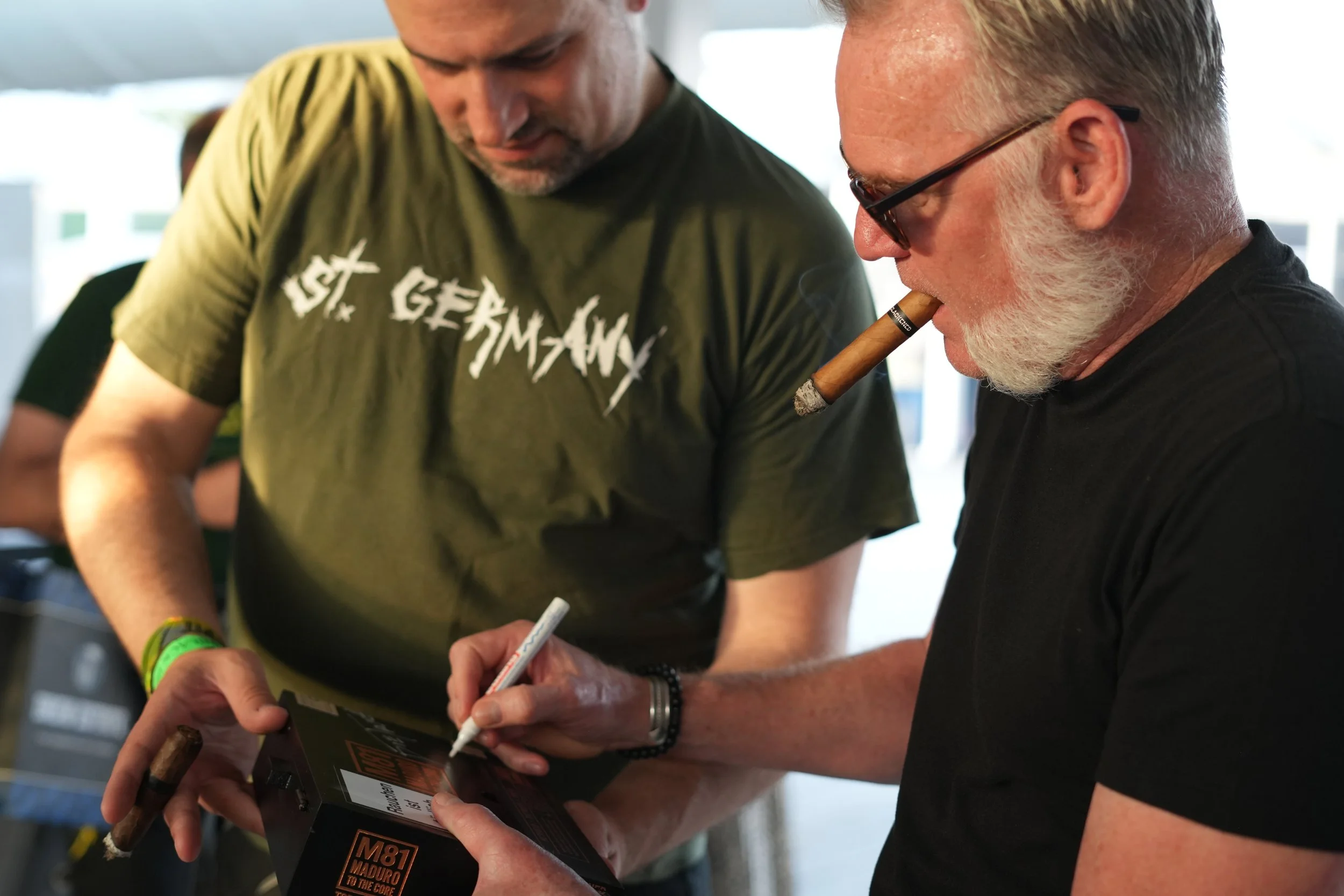 Two men are closely looking at or signing a black box labeled "M81 MADURO TWICE THE CORE." One is wearing a green T-shirt and a green wristband, while the other has gray hair, glasses, and is smoking a cigar.