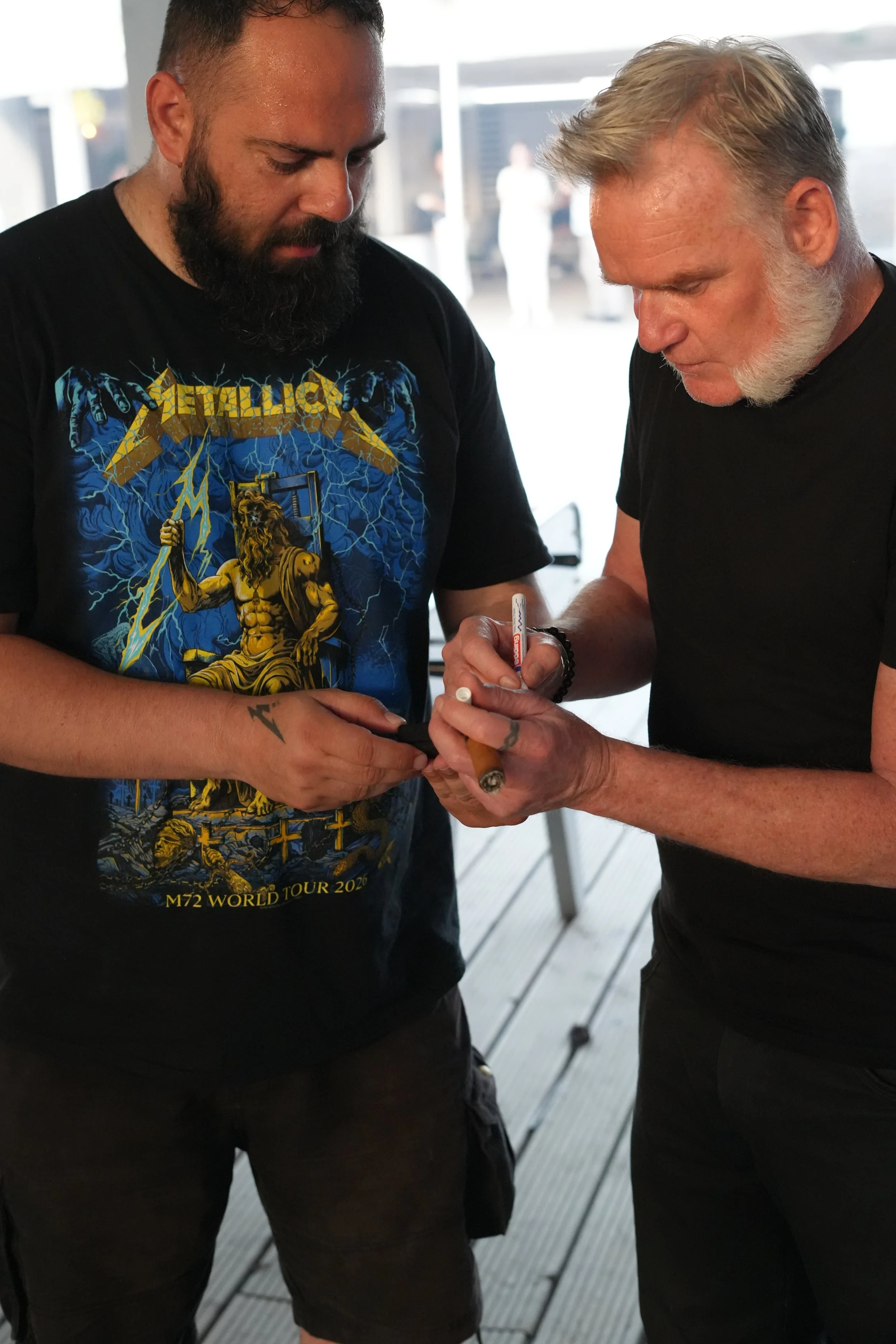 Two men signing an autograph together, one wearing a black Metallica T-shirt and the other holding a marker.