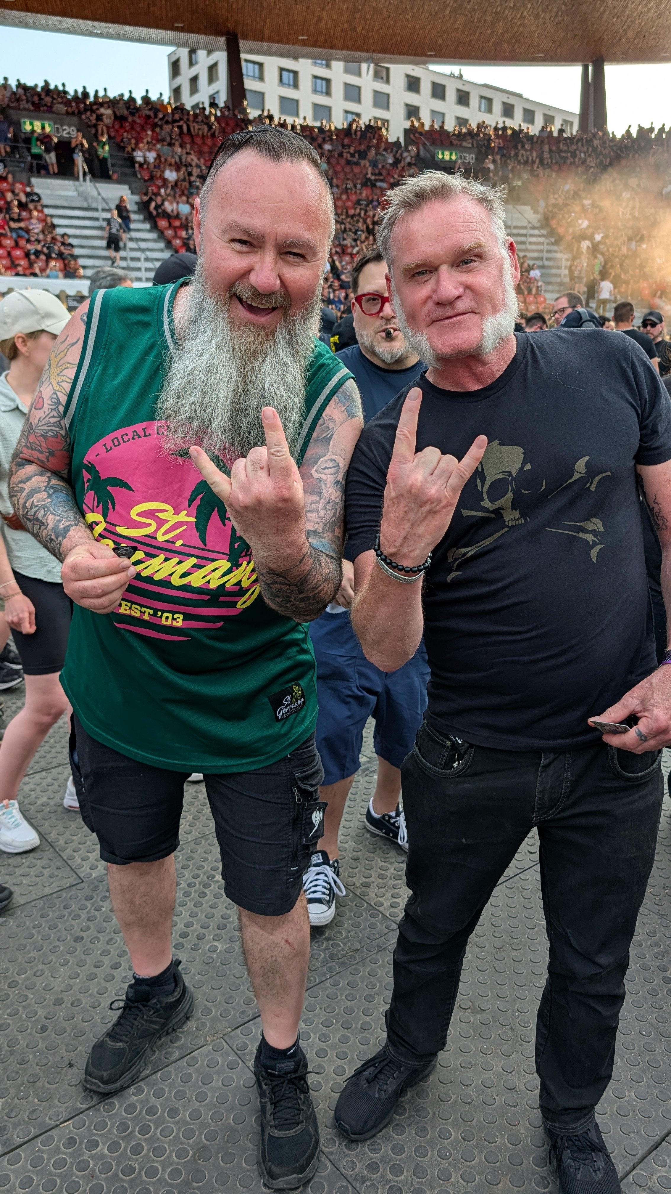 Two men smiling and making the rock and roll hand gesture at a Metallica concert with a crowd in the background.