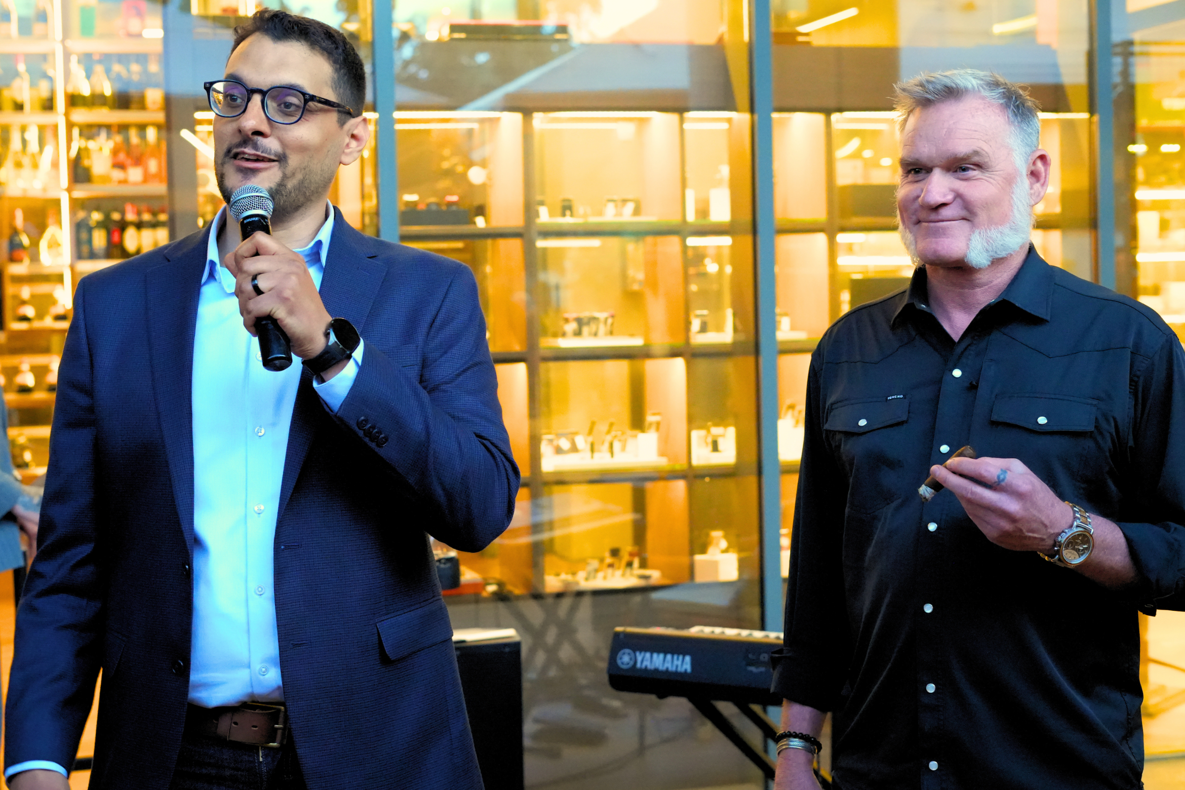 Two men stand indoors in front of a glass wall with warm lighting. The man on the left is wearing glasses, a blue suit, and a white shirt, and is holding a microphone. The man on the right has gray hair and a beard, is wearing a black shirt, and is h