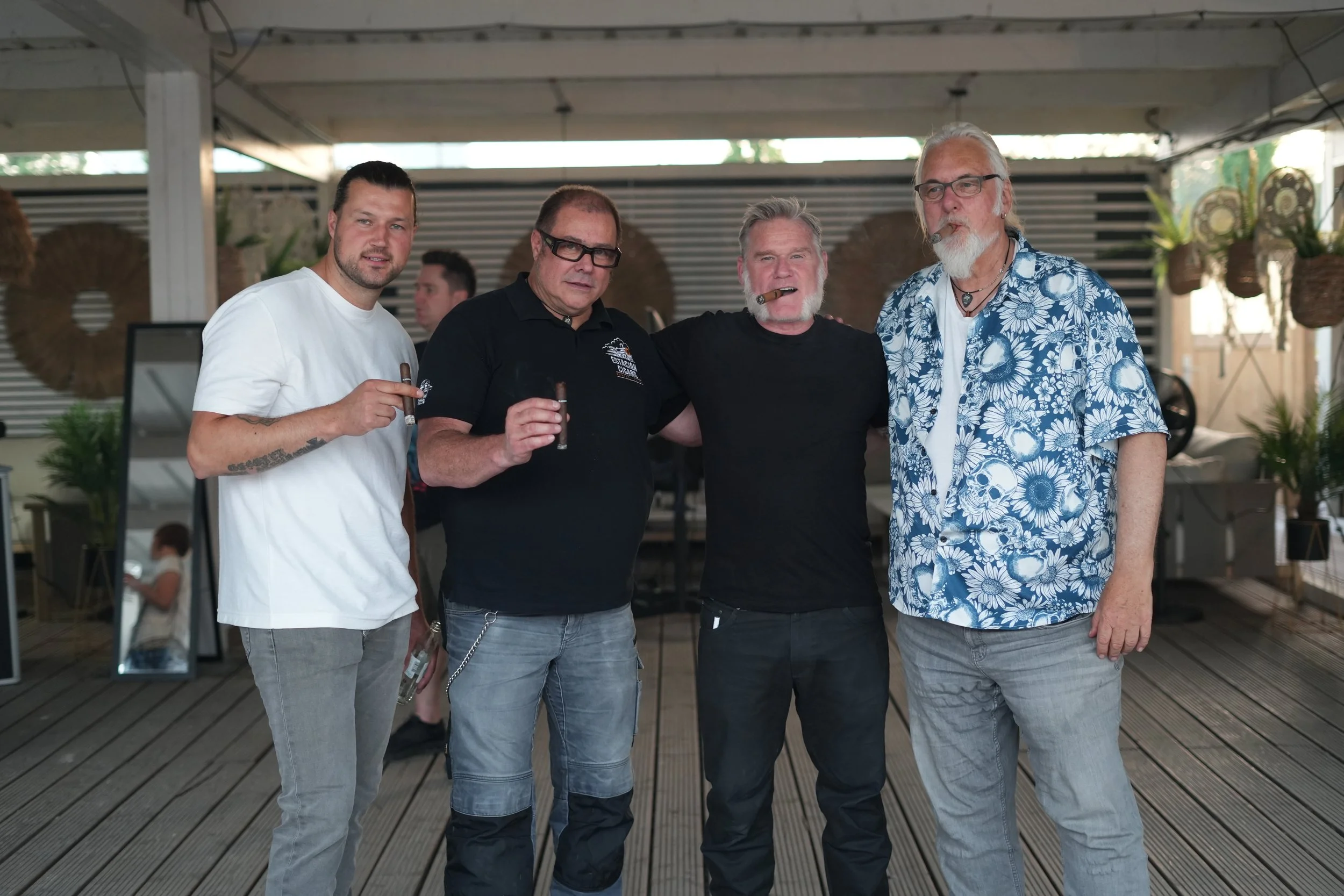 Four men posing together outdoors under a pergola, some holding cigars, with a decorative wall and hanging baskets in the background.
