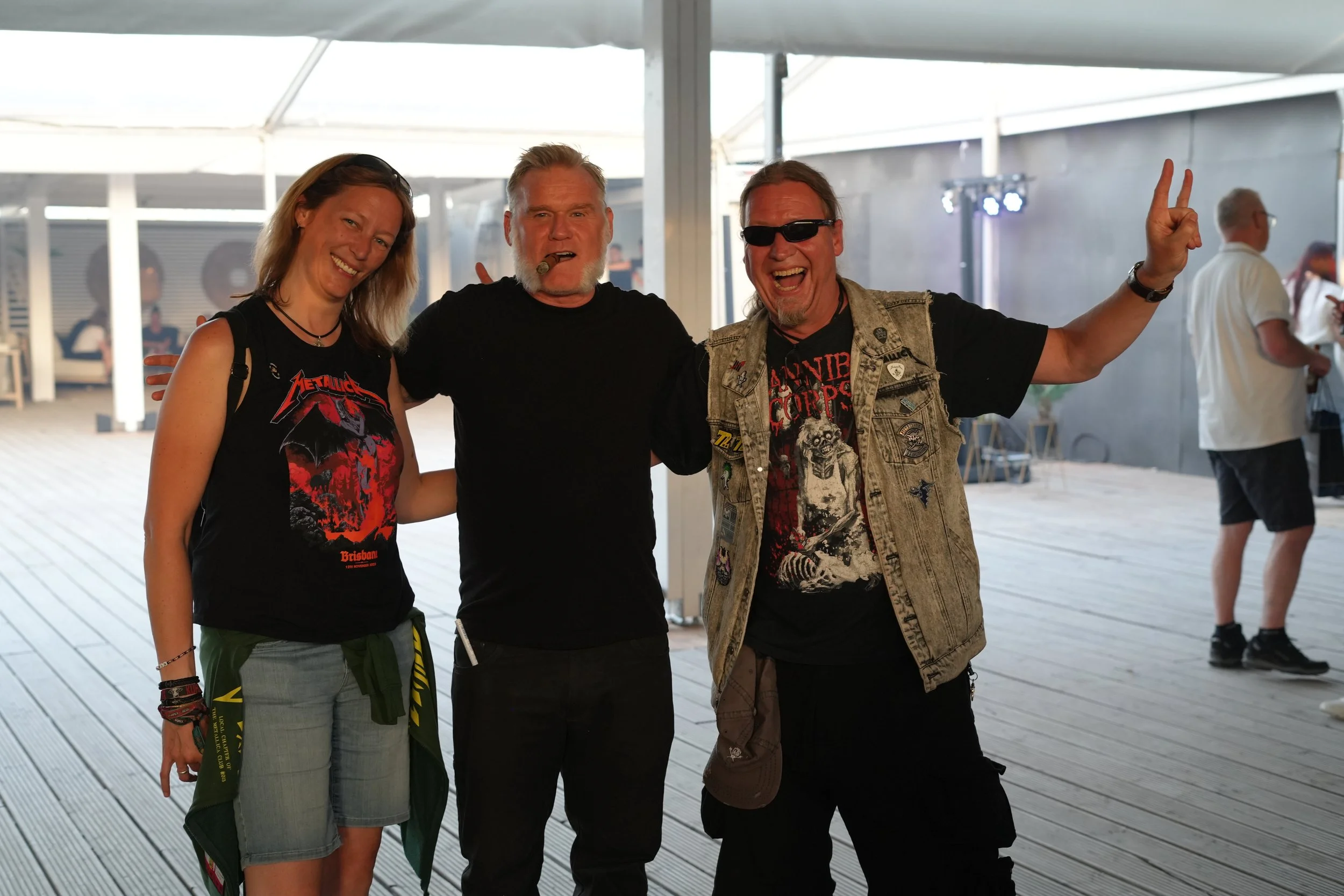 Three people smiling and posing for a photo, standing outdoors under a tent. The person on the left is a woman with light brown hair, wearing a black Metallica shirt. The middle person is a man with gray hair and beard, smoking a cigar, wearing a bla