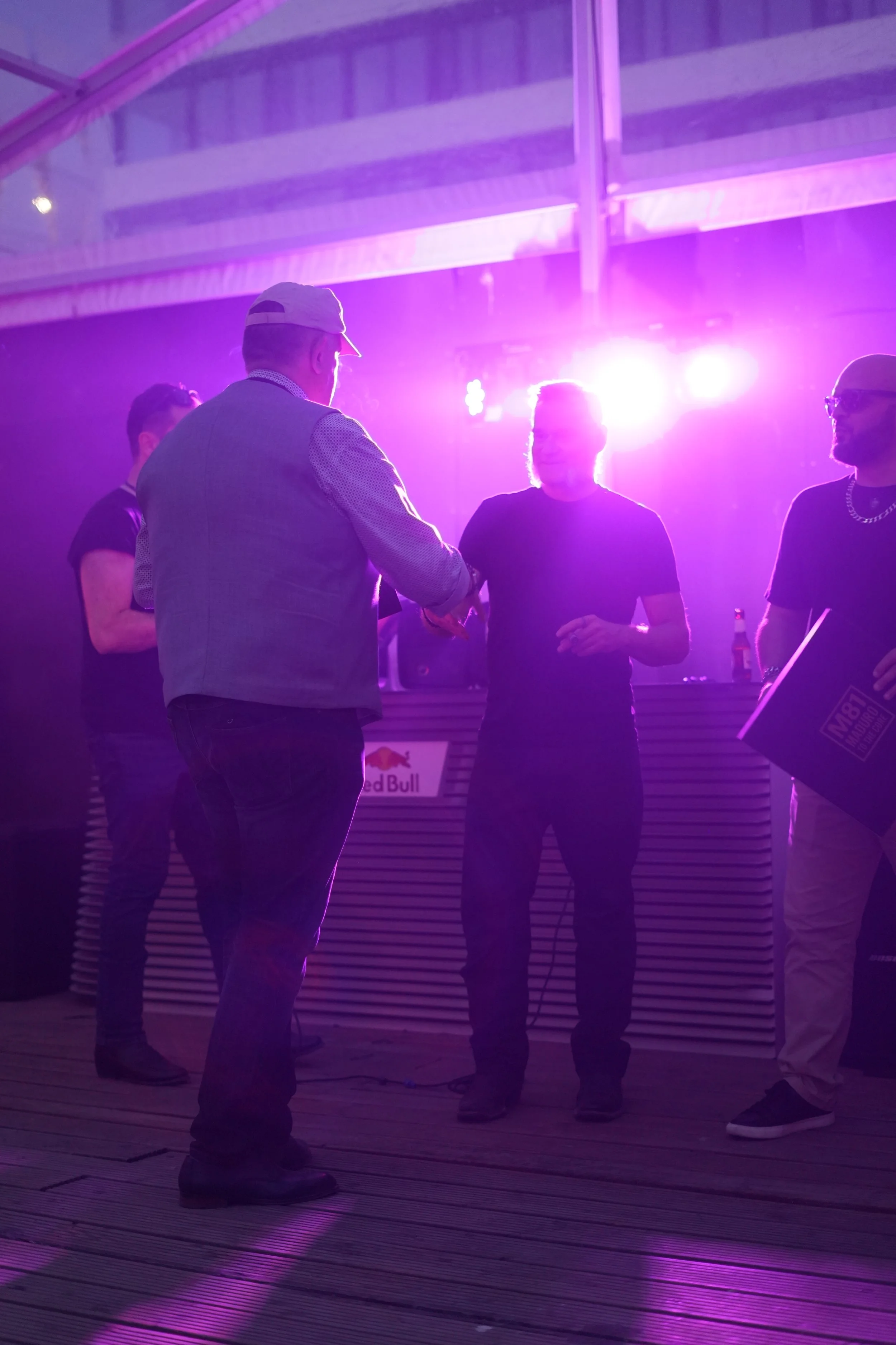 Four men are standing in a circle under purple lighting in an indoor space with wooden flooring and ceiling beams. One man is wearing a hat and glasses, another has a chain necklace, and the others are dressed casually. They seem to be having a conve