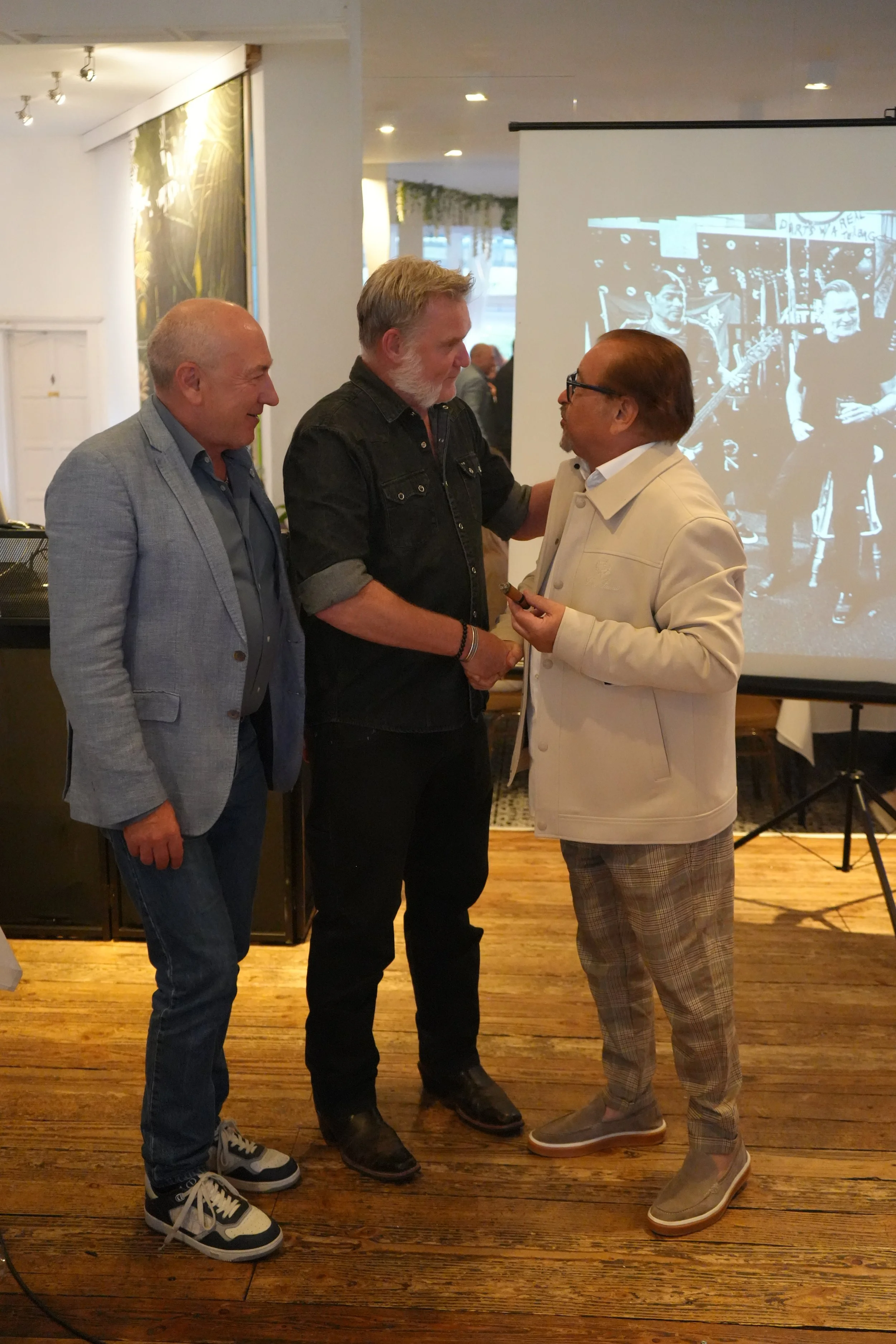 Three men standing indoors, shaking hands and chatting at a social event. One man is wearing a light grey blazer and jeans, another in a black denim shirt and dark jeans, and the third in a white jacket with plaid pants. There is a large black-and-wh