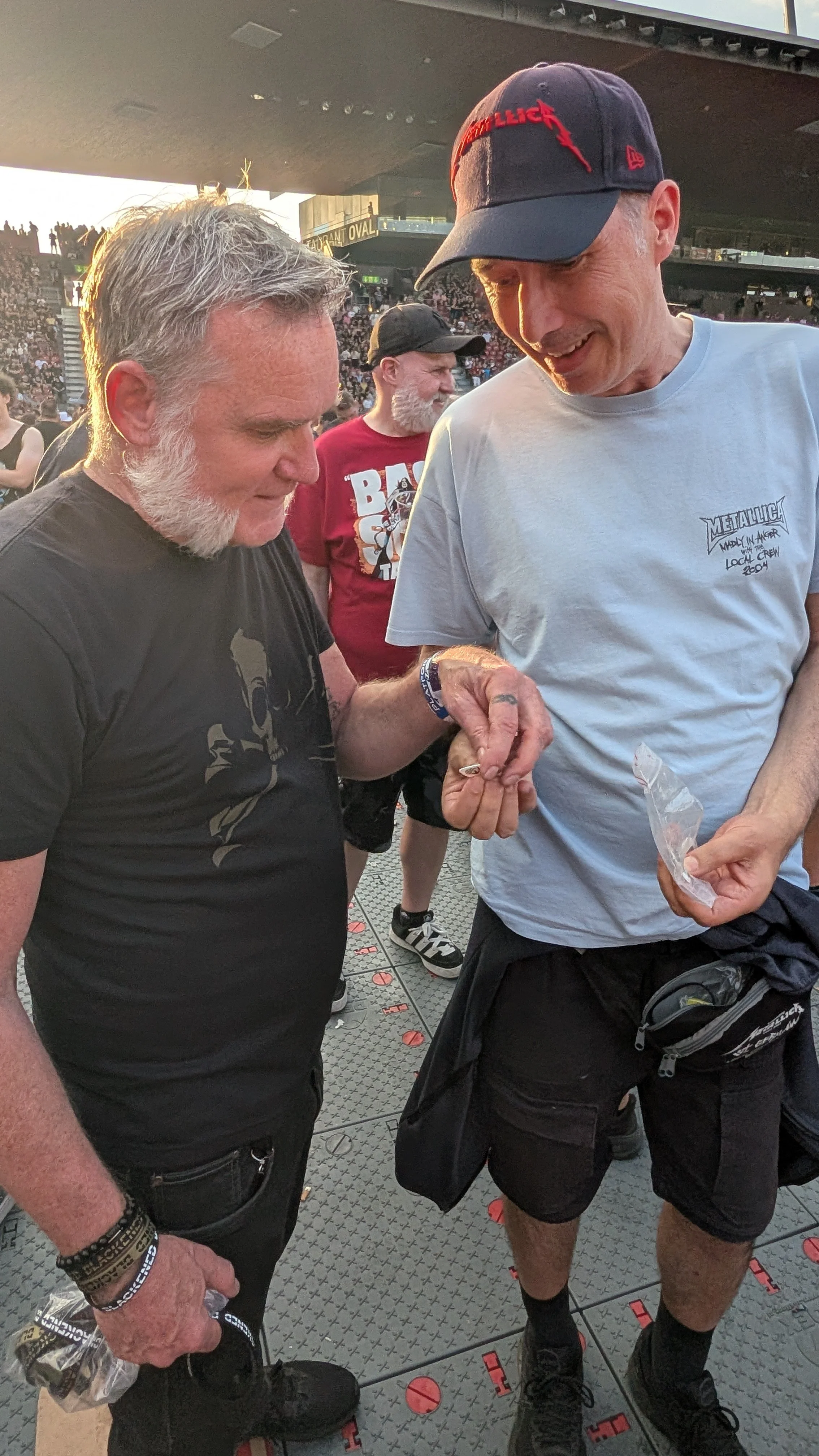 Two men are at a Metallica concert, with a crowd behind them. They are interacting, with the older man holding a small object, and the younger man holding a plastic bag. The event appears to be outdoors in a stadium or large venue.