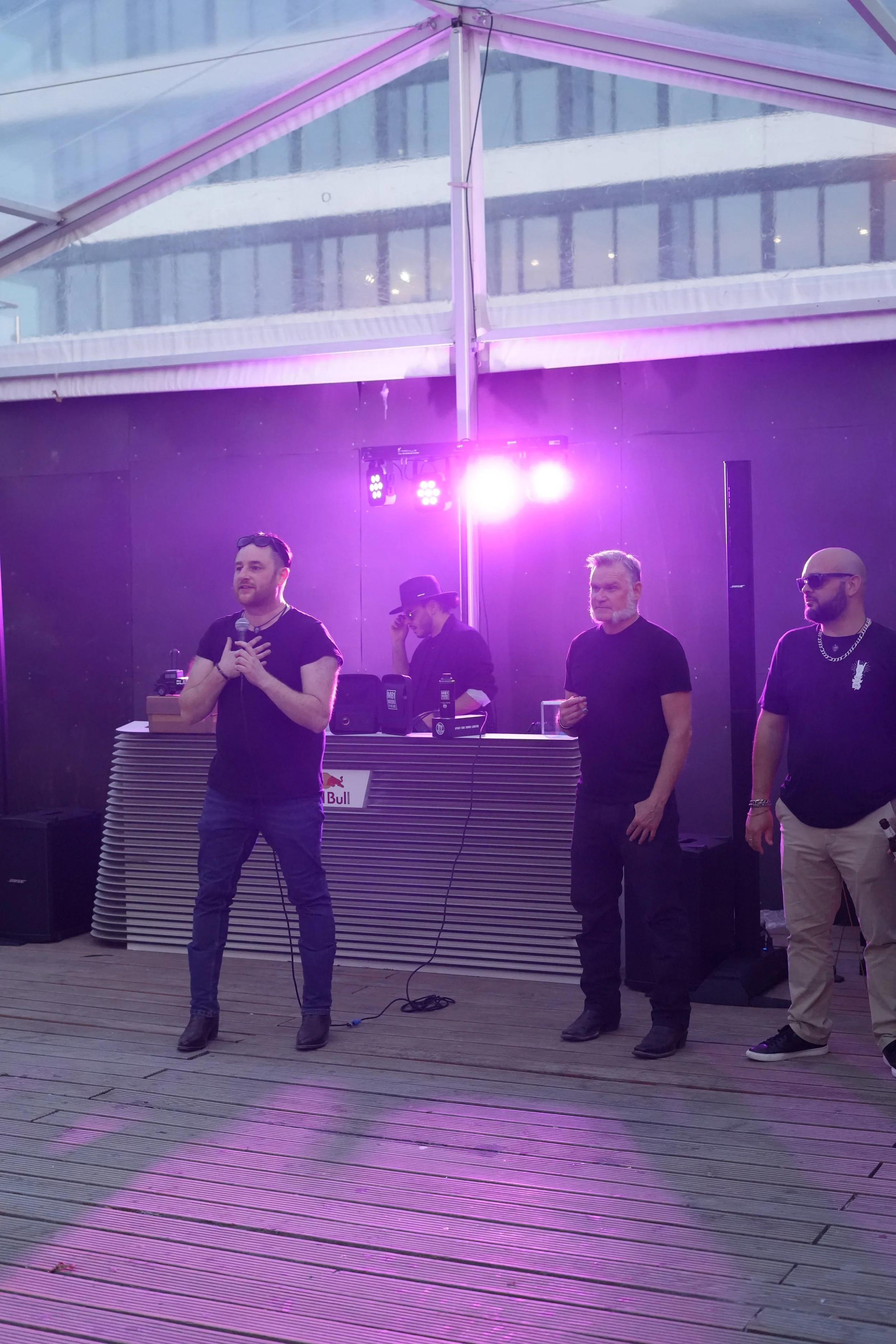 Four men standing on a wooden deck under a tent with purple lighting. One man is holding a microphone, and two are wearing sunglasses. There is DJ equipment on a table behind them with a Red Bull logo.