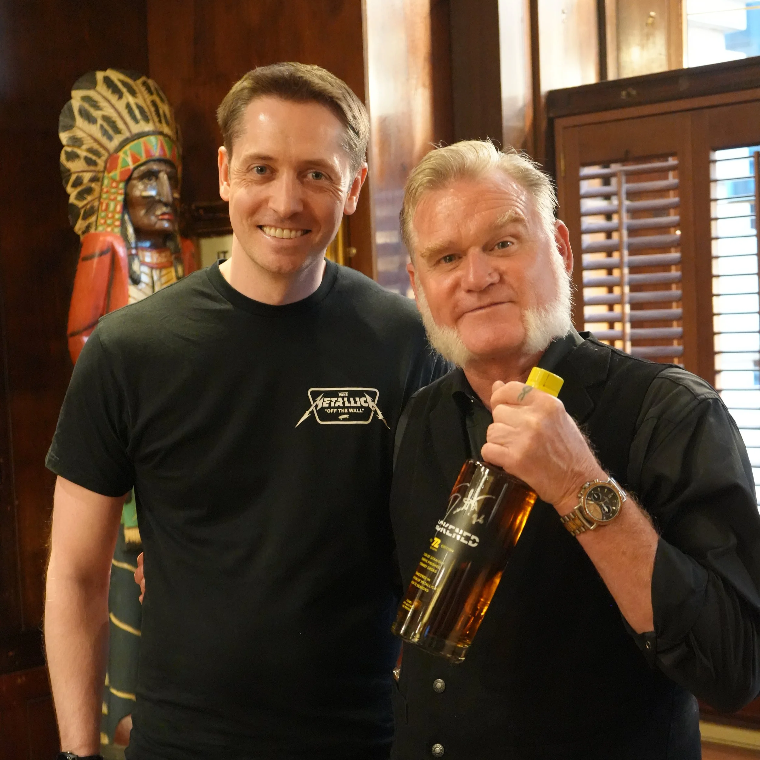 Two men smiling, one holding a bottle of Blackened whiskey, inside a wooden-paneled room with a decorative tribal mask on the wall and wooden window shutters.