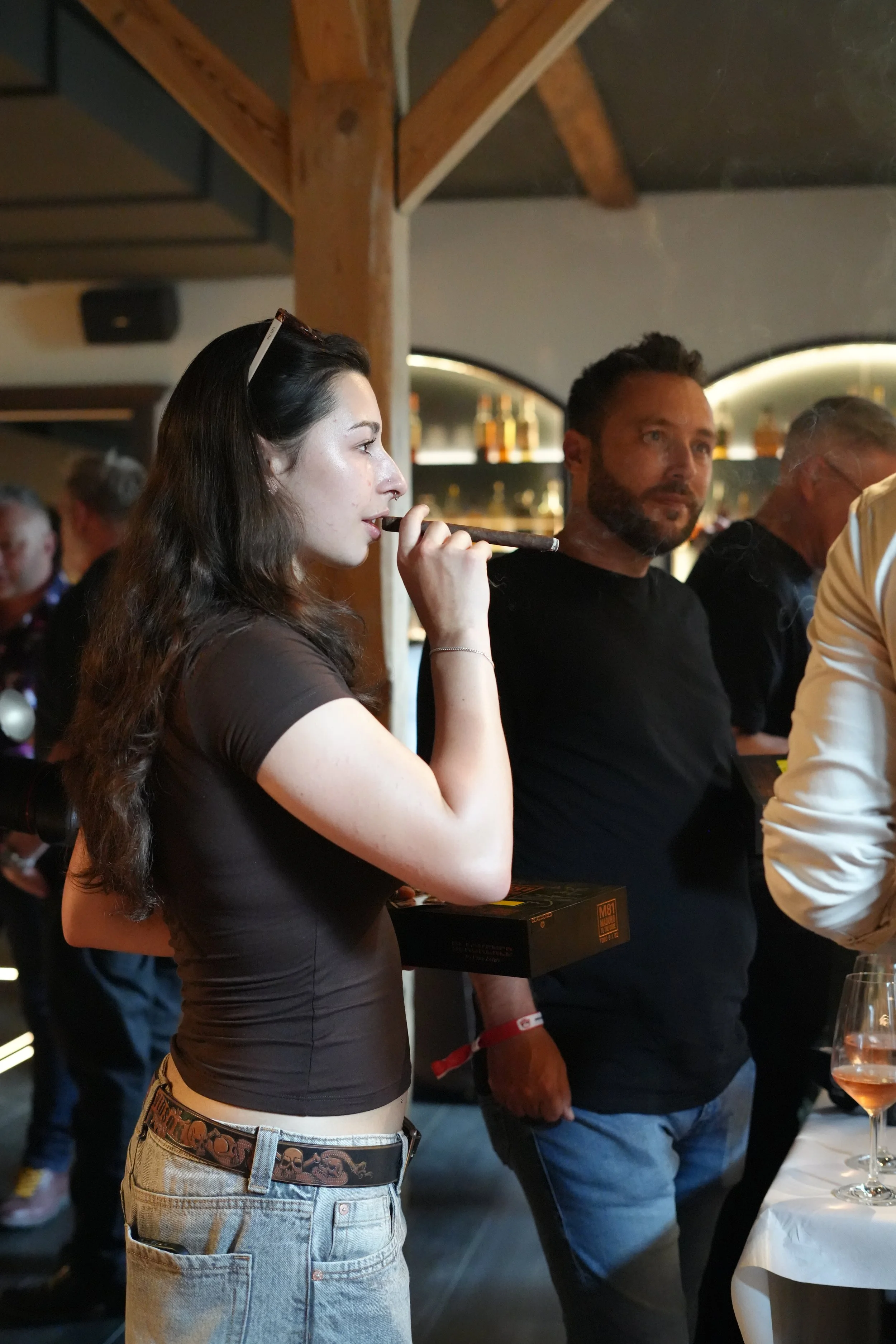 A young woman with long brown hair, wearing a brown shirt and light-colored jeans with a skull belt, is standing holding a cigar. She is near a man with a beard and black shirt, who is looking at her, while other people are in the background in a bar