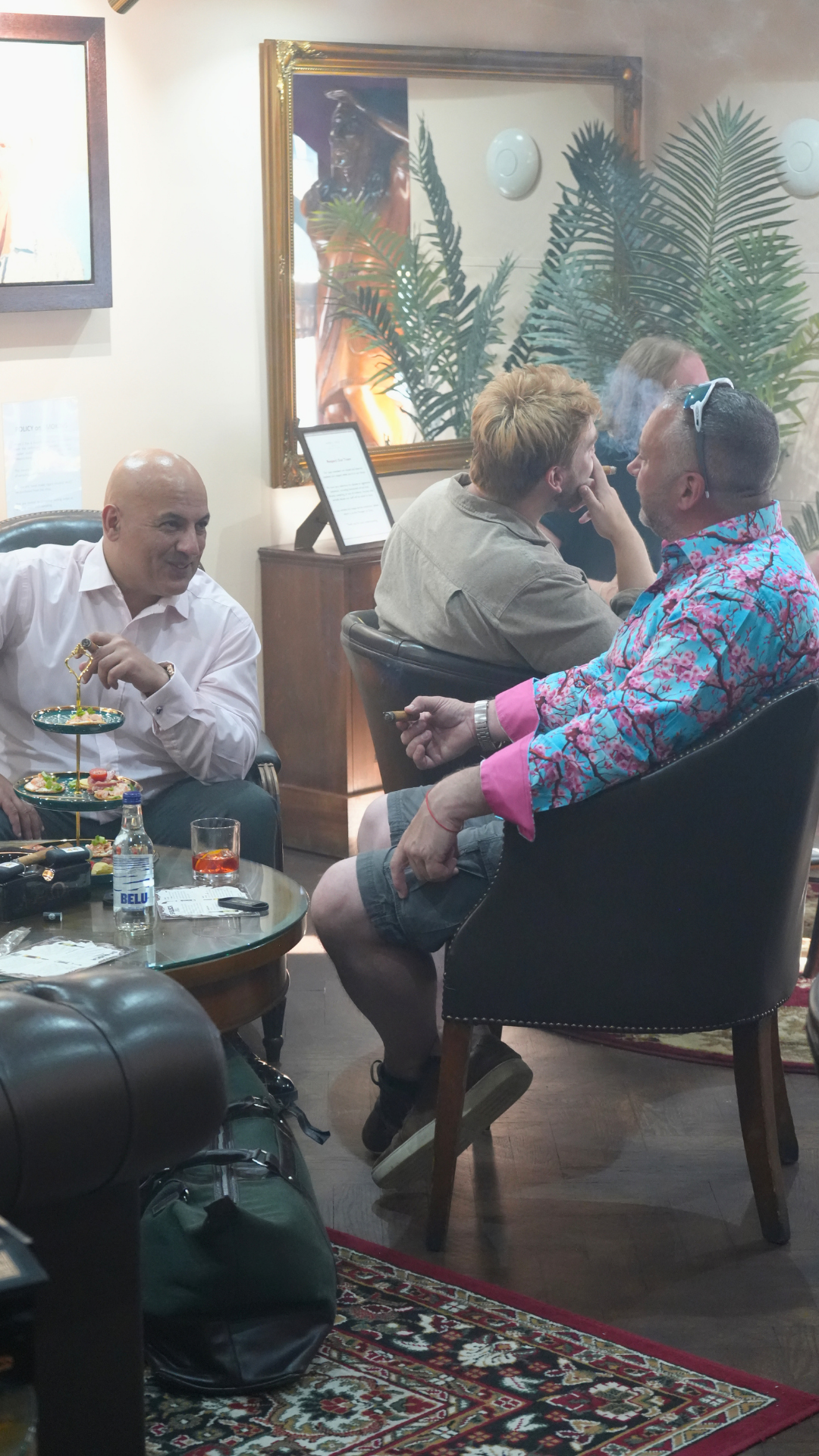 Group of men socializing in a room with a mirror, plants, and framed pictures on the wall. One man is holding a cigarette, both are sitting in chairs, and there is a table with snacks, drinks, and a backpack in front of them.