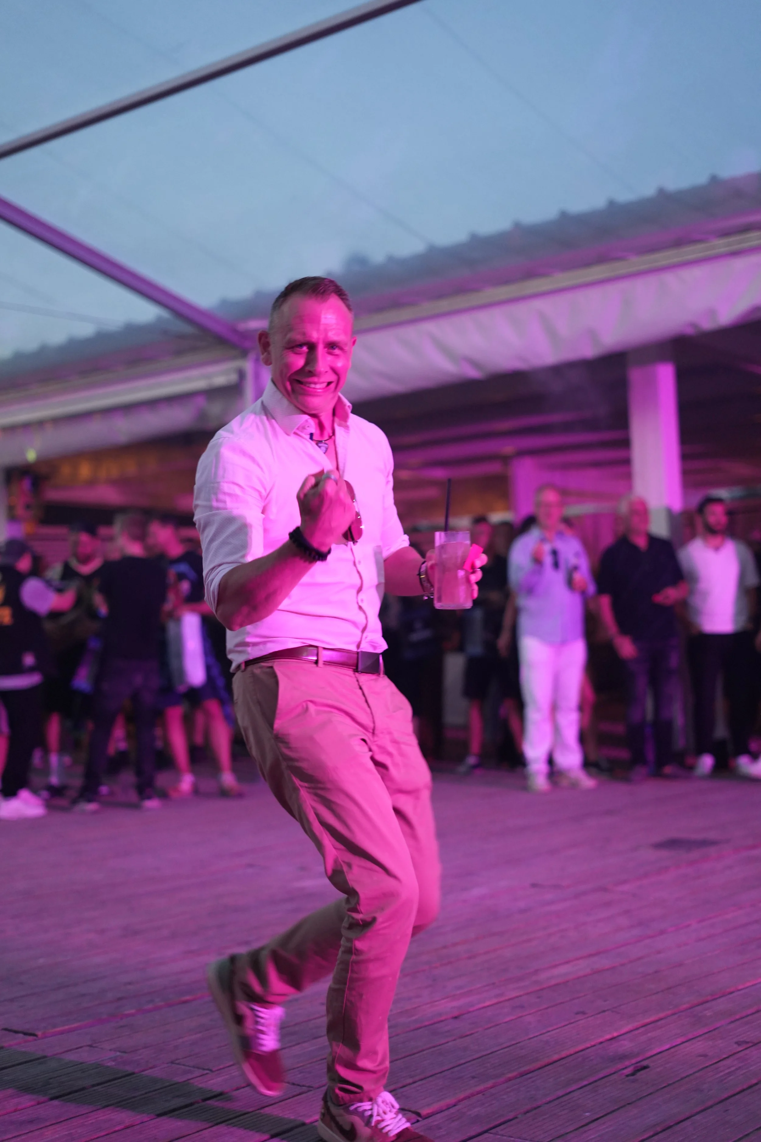 A man dancing at a lively event under a tent with pink lighting, wearing a white shirt, beige pants, and sneakers, with a drink in his hand and a big smile.