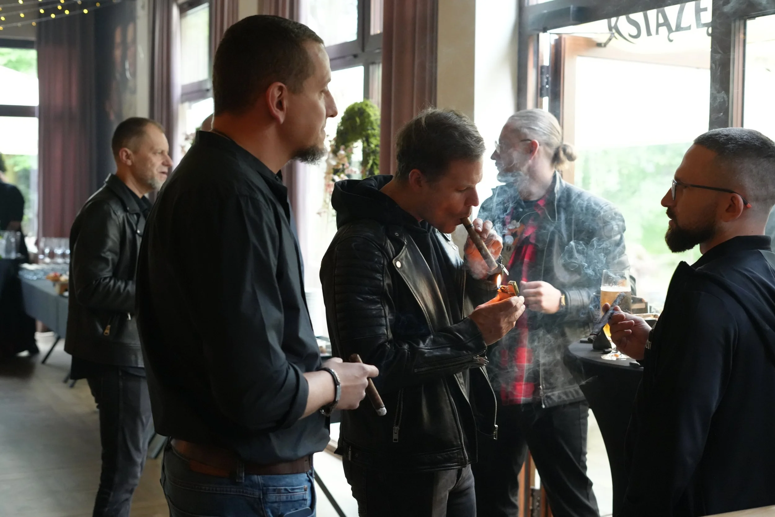 Group of men socializing at a gathering, with some smoking cigars and drinking, indoors near windows with natural light.