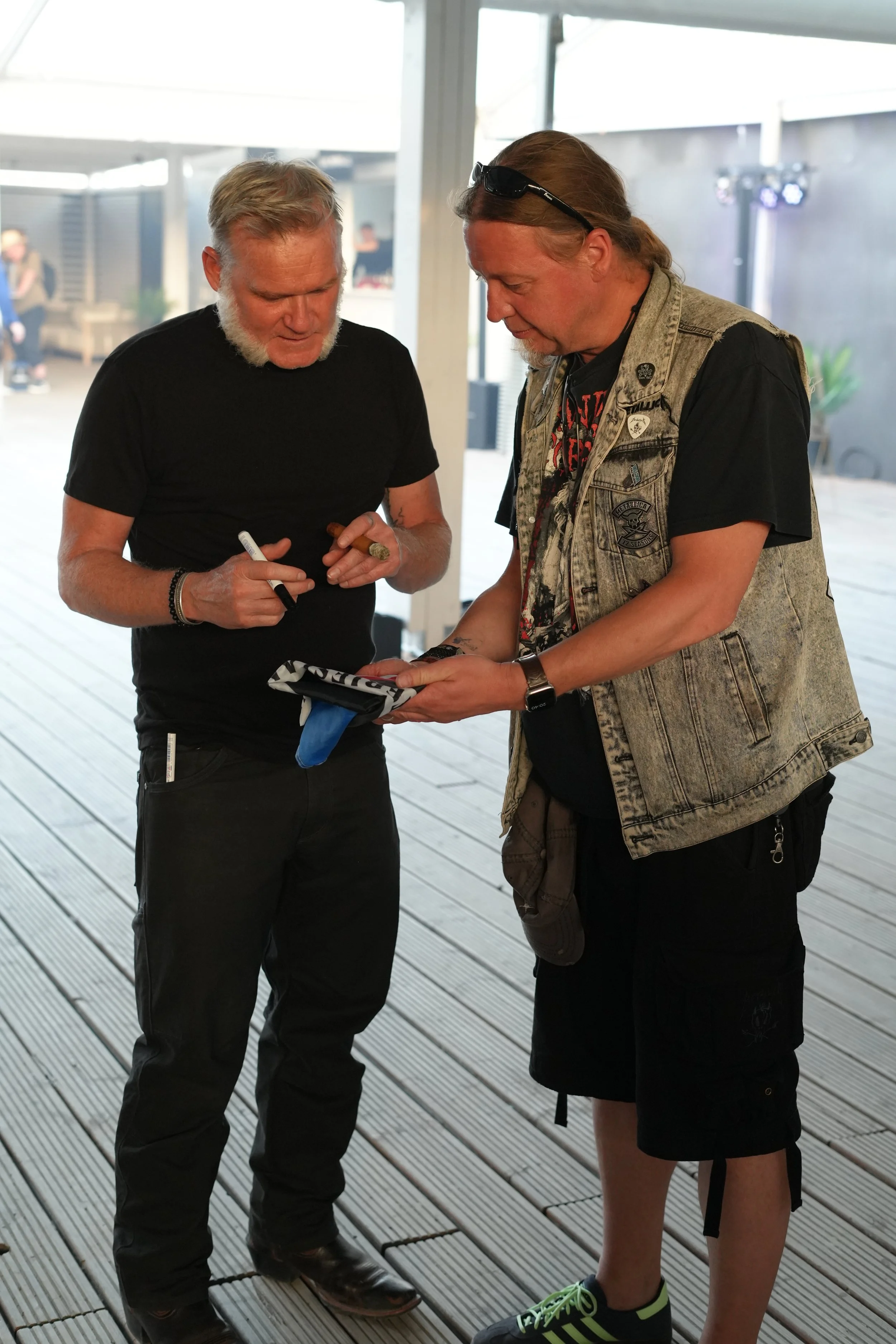 Two men standing on a wooden deck, looking at something in one man's hands, with one holding a cigarette and the other wearing sunglasses on his head.