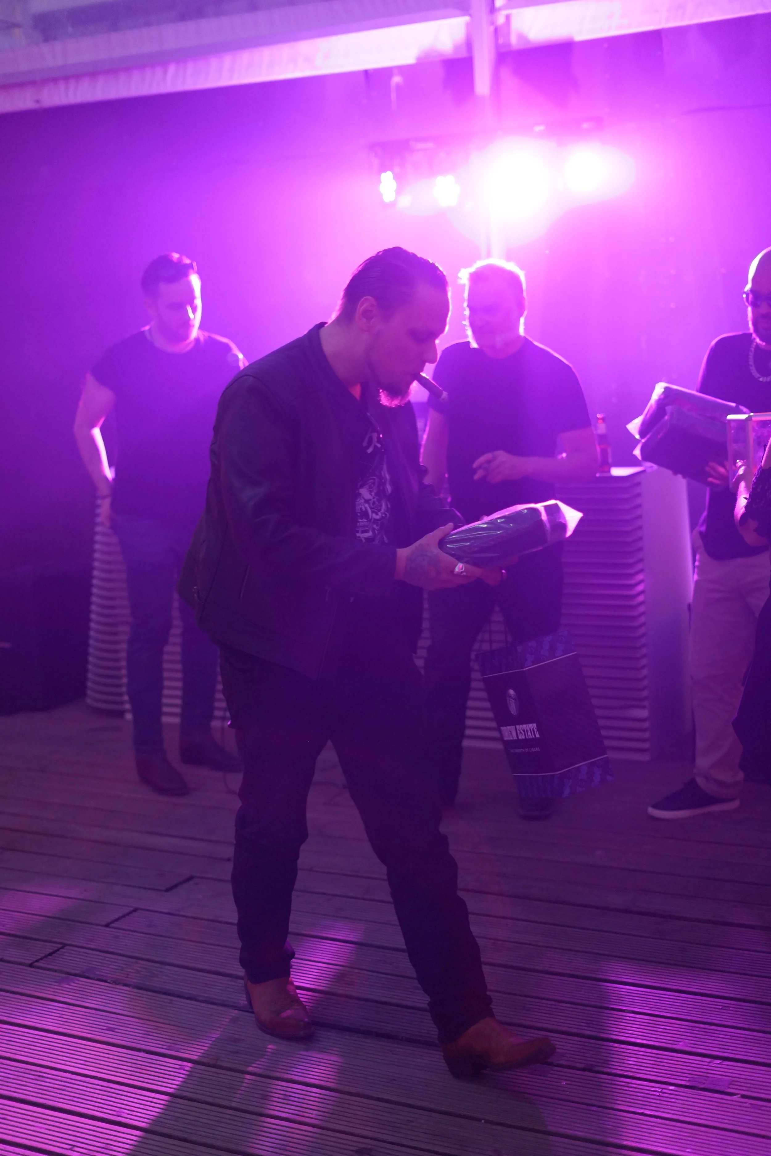 Group of people in a dimly lit room with pink and purple lighting, some holding books or magazines, standing on a wooden floor.