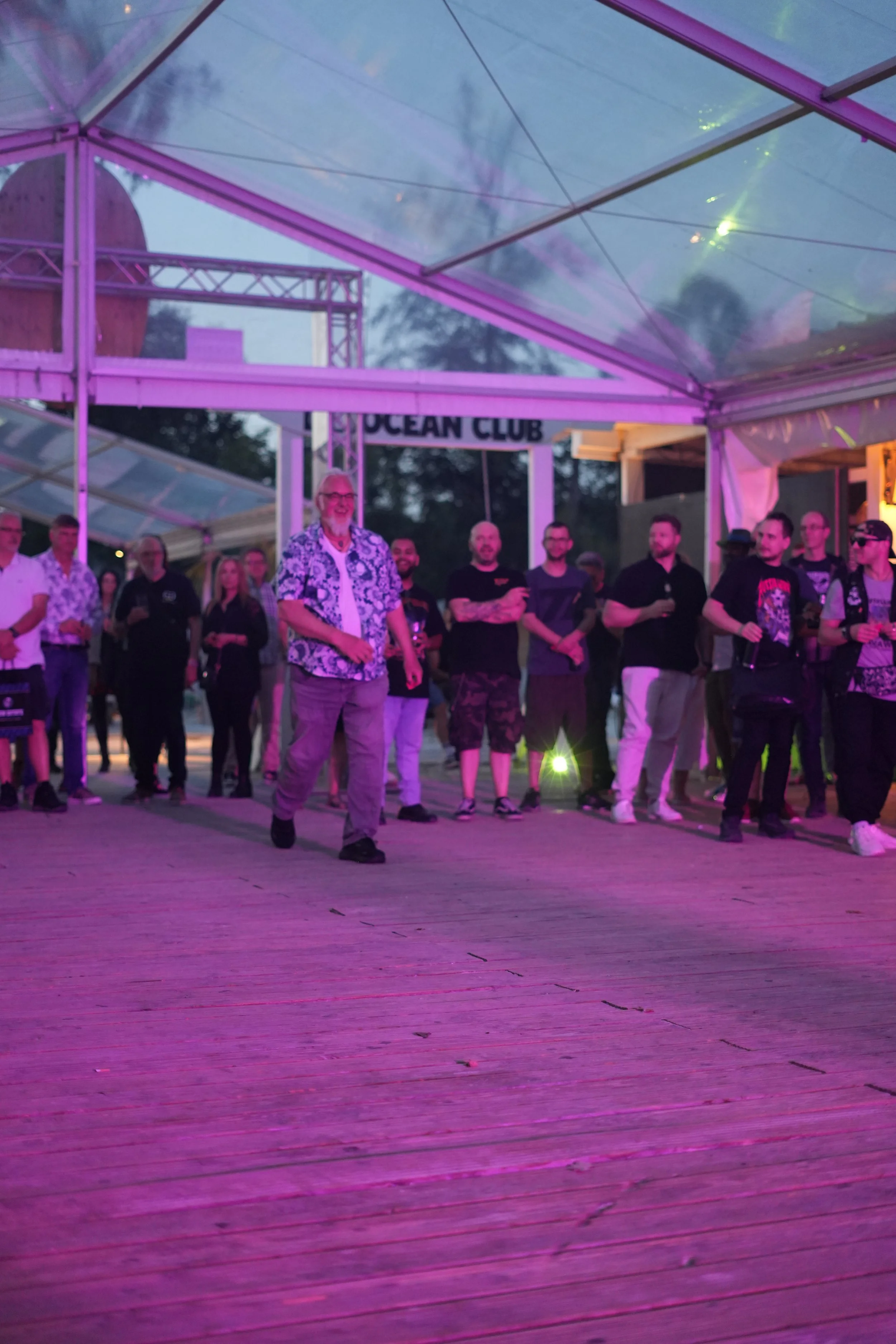 People gathered at an outdoor event under a transparent tent with purple lighting, with a sign reading 'OCEAN CLUB' in the background.