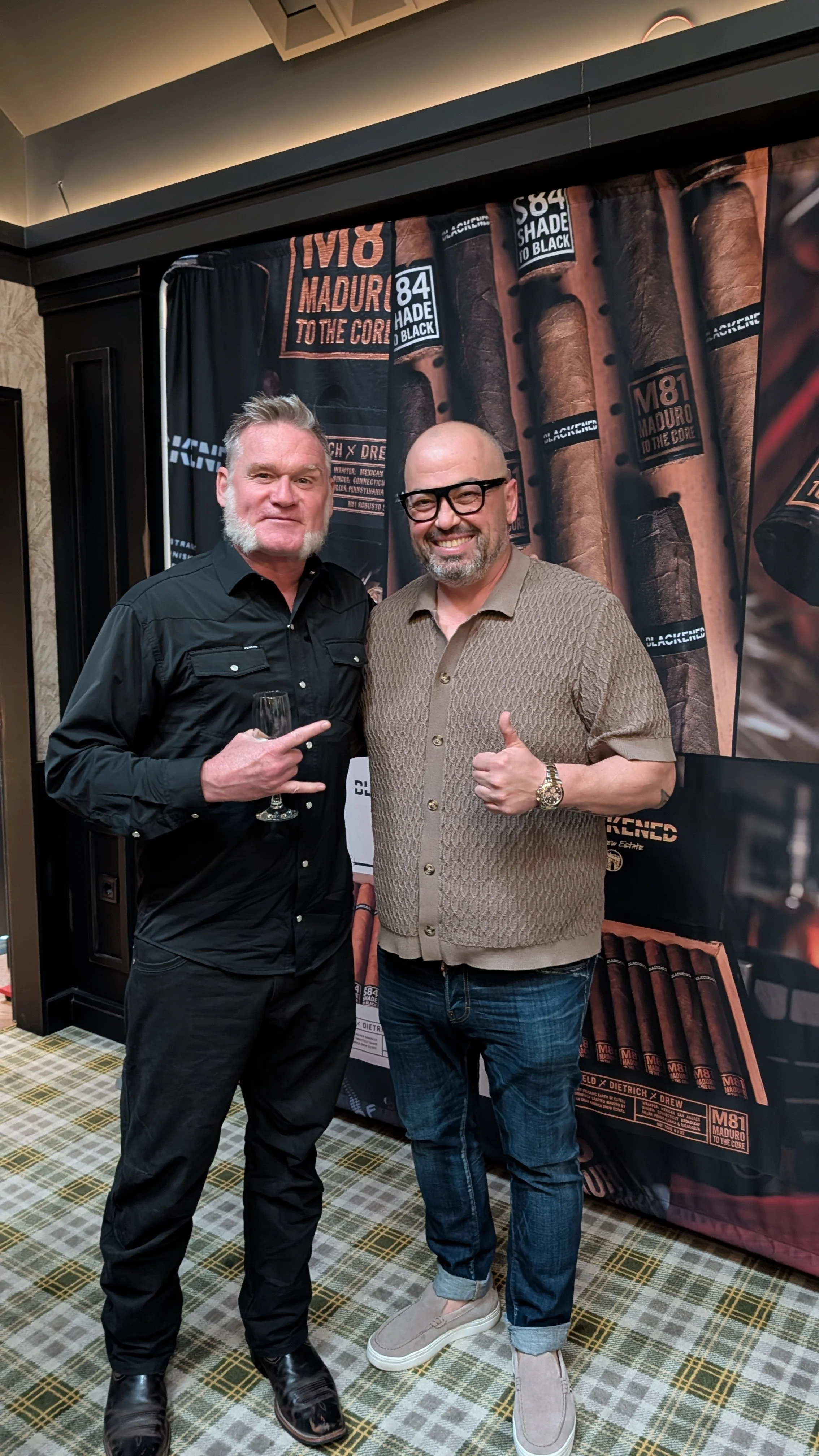 Two men standing in front of a large banner featuring cigars, smiling at the camera. One man is wearing a black shirt and the other a beige patterned shirt and jeans.
