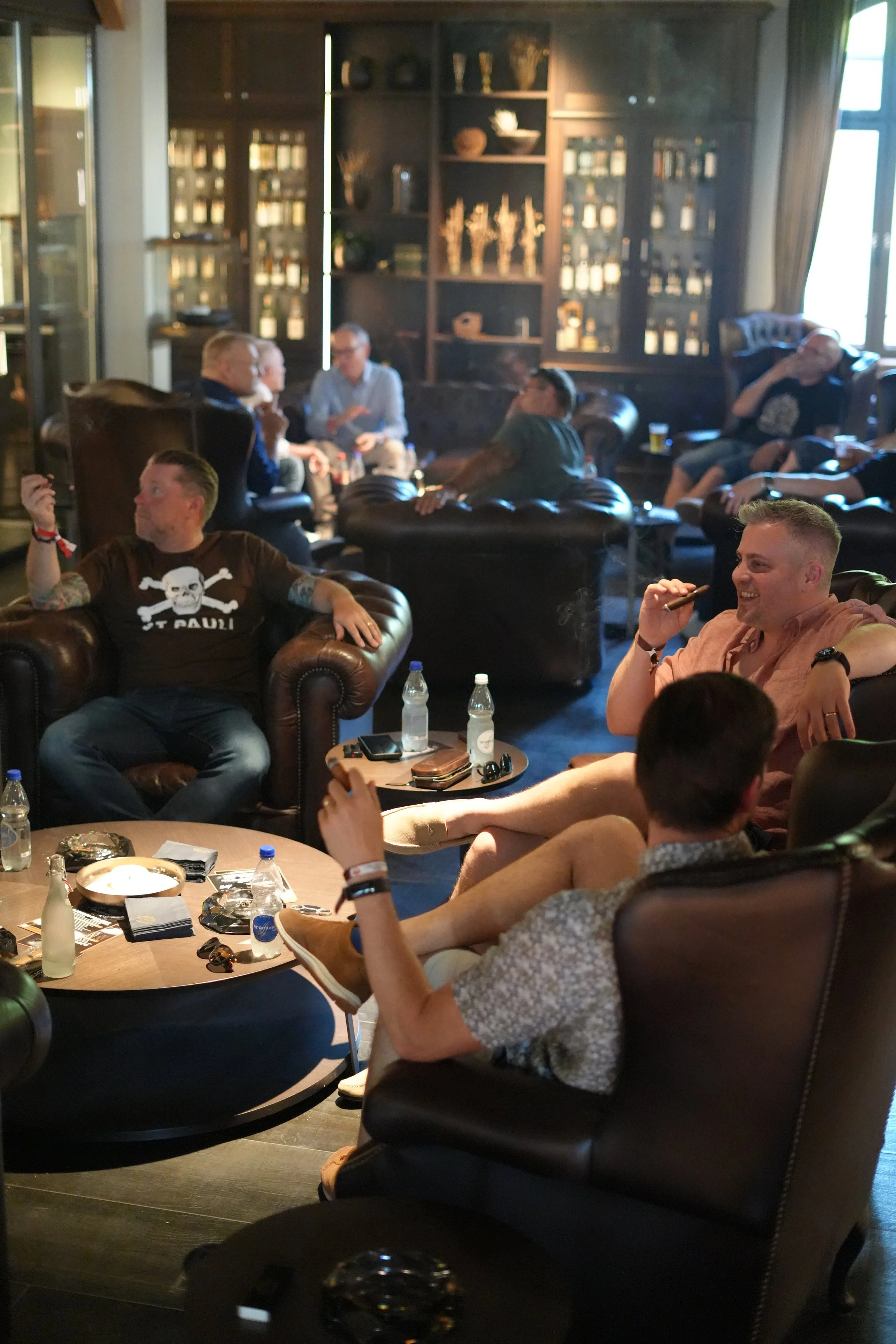 People sitting and chatting in a cozy lounge with dark wooden bookshelves filled with bottles and decorative items in the background. Some individuals are holding drinks and socializing around tables with snacks and beverages.