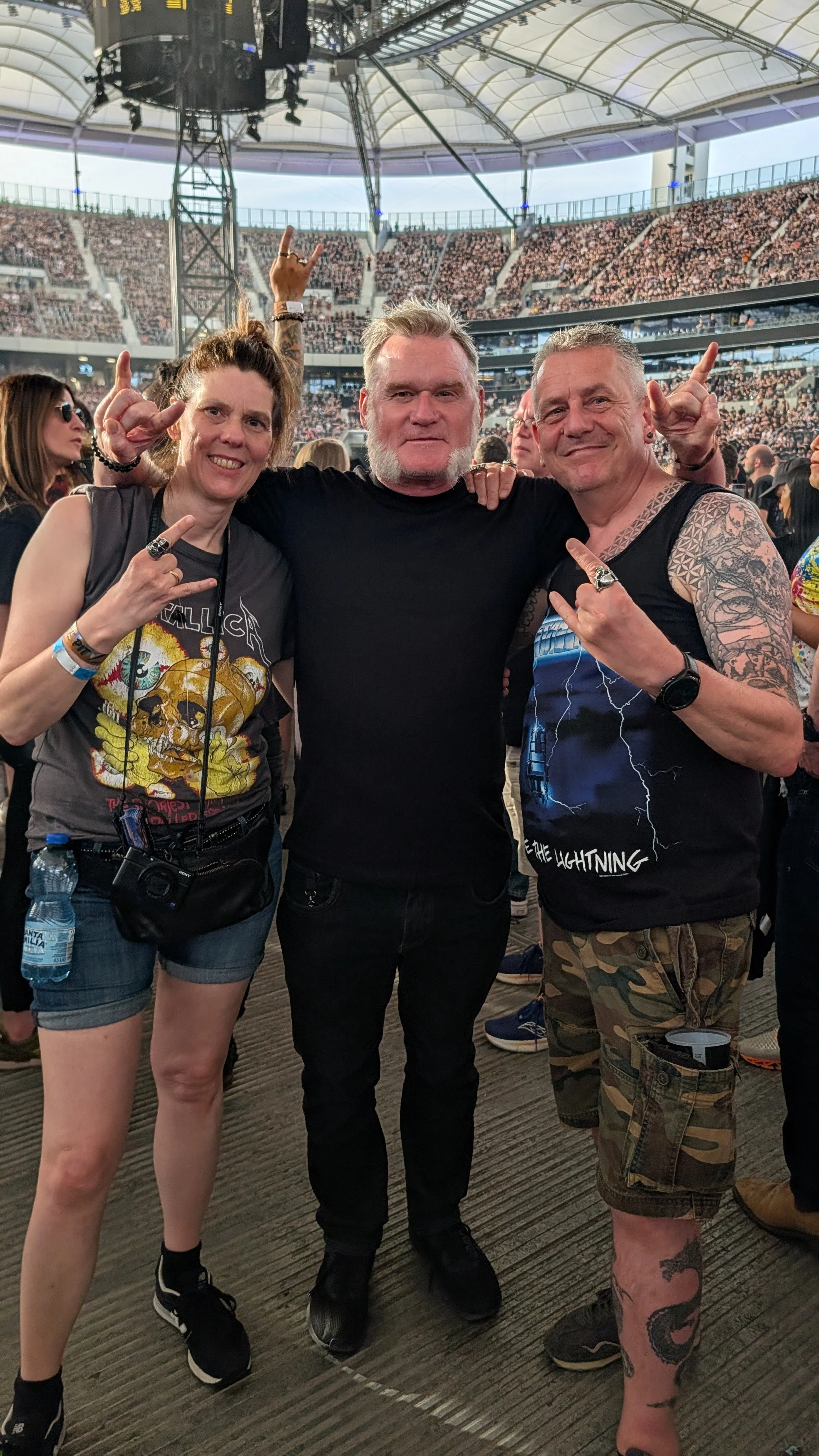 Three people at a concert or music event posing and making rock hand gestures, with a large audience and stadium architecture in the background.