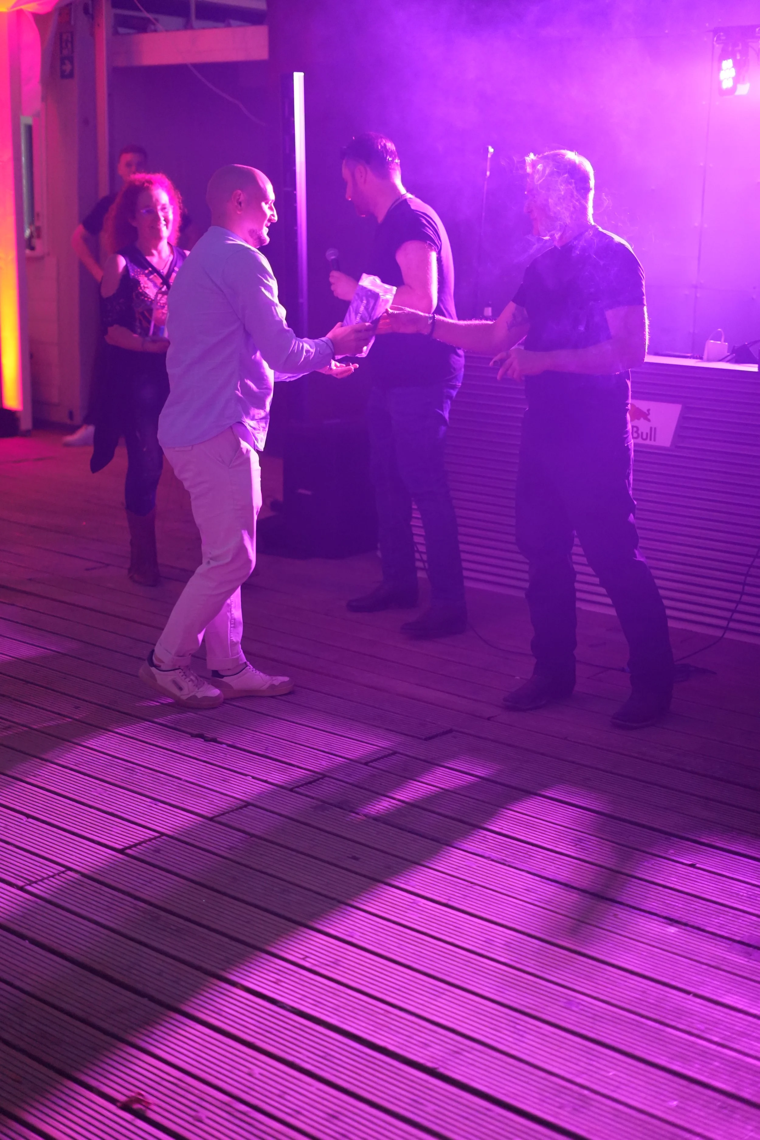 Four people are standing on a wooden floor under purple lighting in a nightclub or event space. Two men are exchanging a gift while a woman watches nearby.
