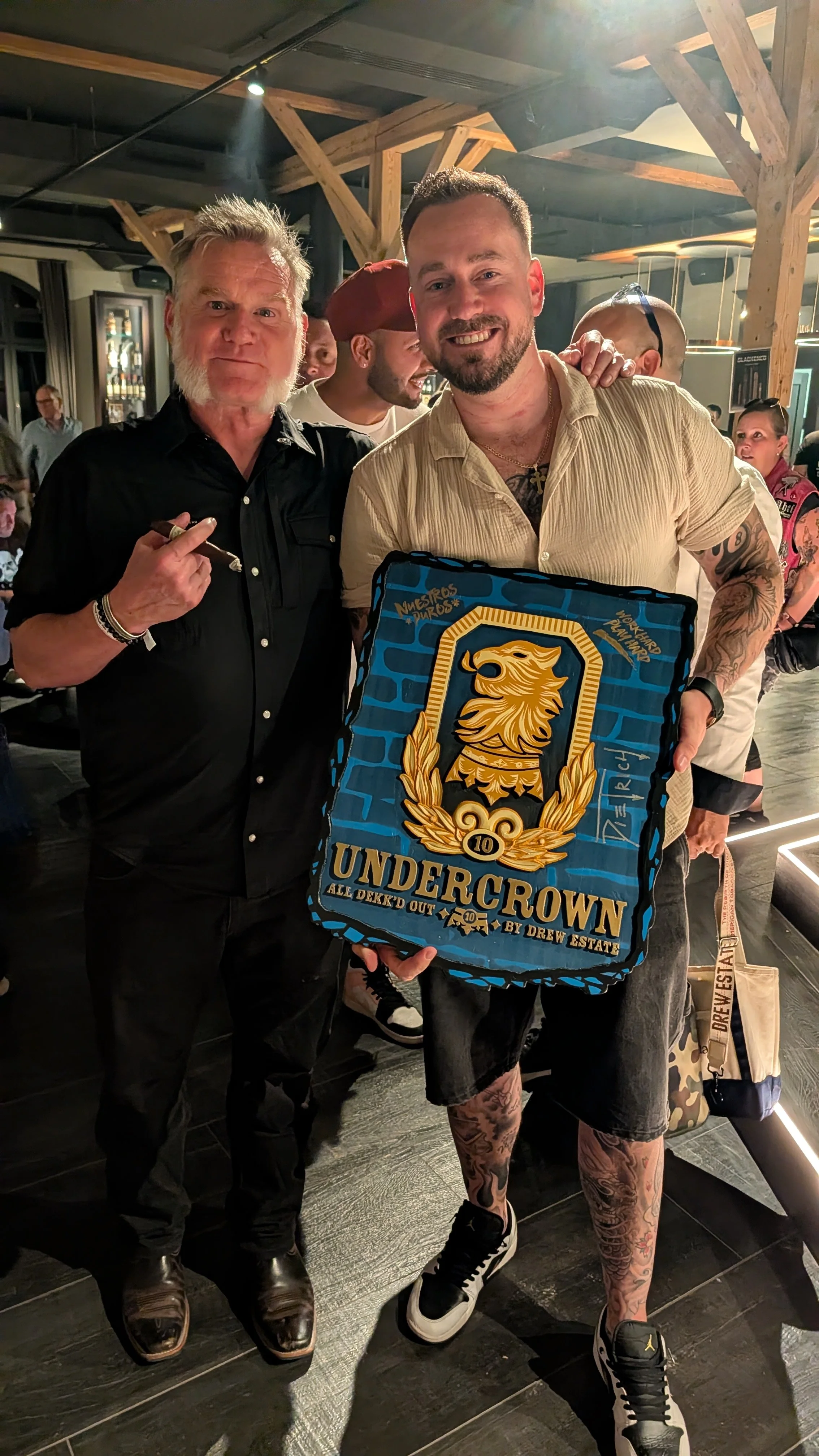 Two men standing in a crowded indoor venue, one with a beard and tattoos holding a large sign with a lion emblem and the word "UNDERCROWN," and the other with gray hair and a cigar pointing at the sign, with several people in the background.