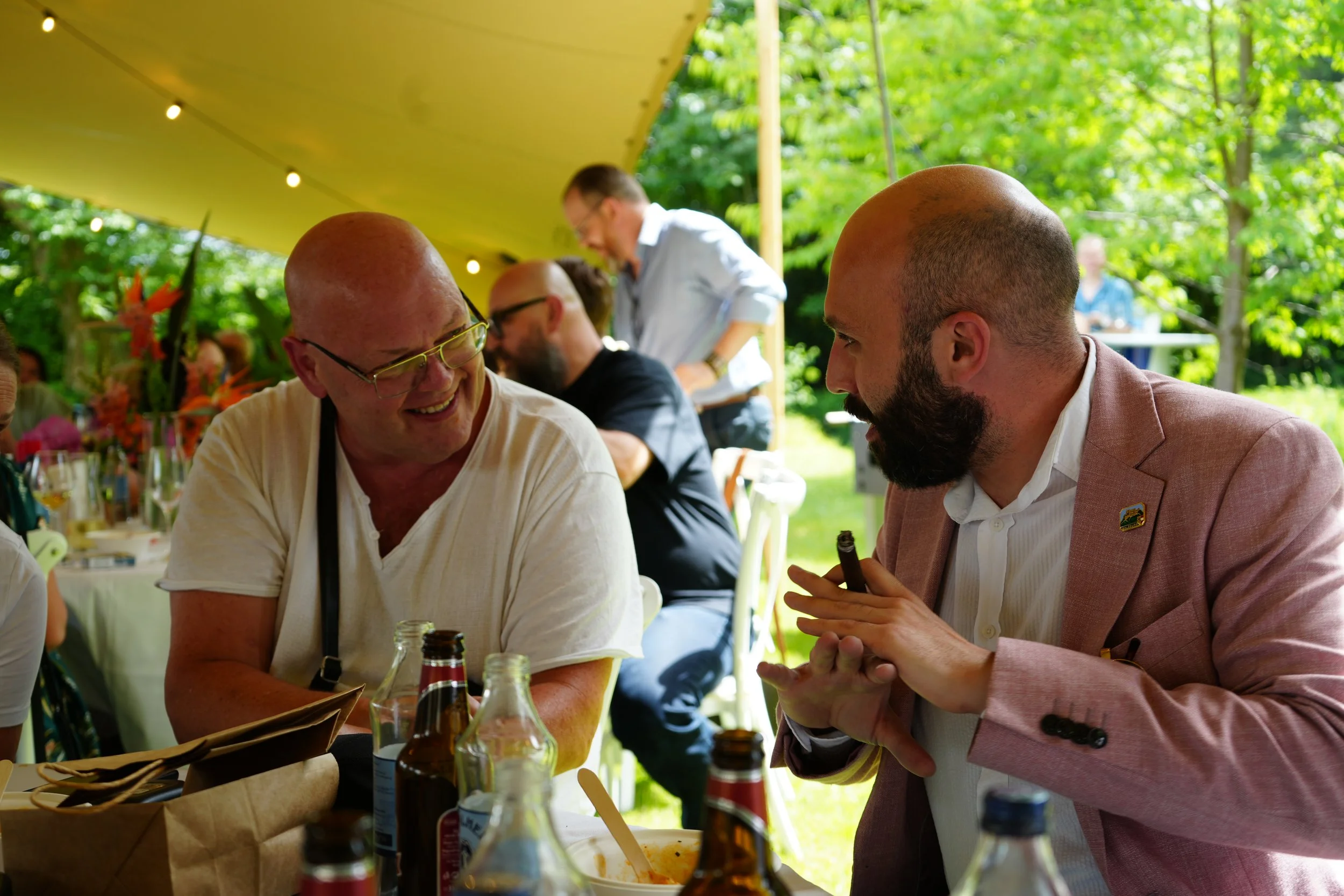 Two men are sitting at a table outdoors, engaging in conversation. One is bald with glasses and wearing a white shirt, and the other has a beard, is bald, and is wearing a pink suit jacket. They are under a canopy with string lights and surrounded by