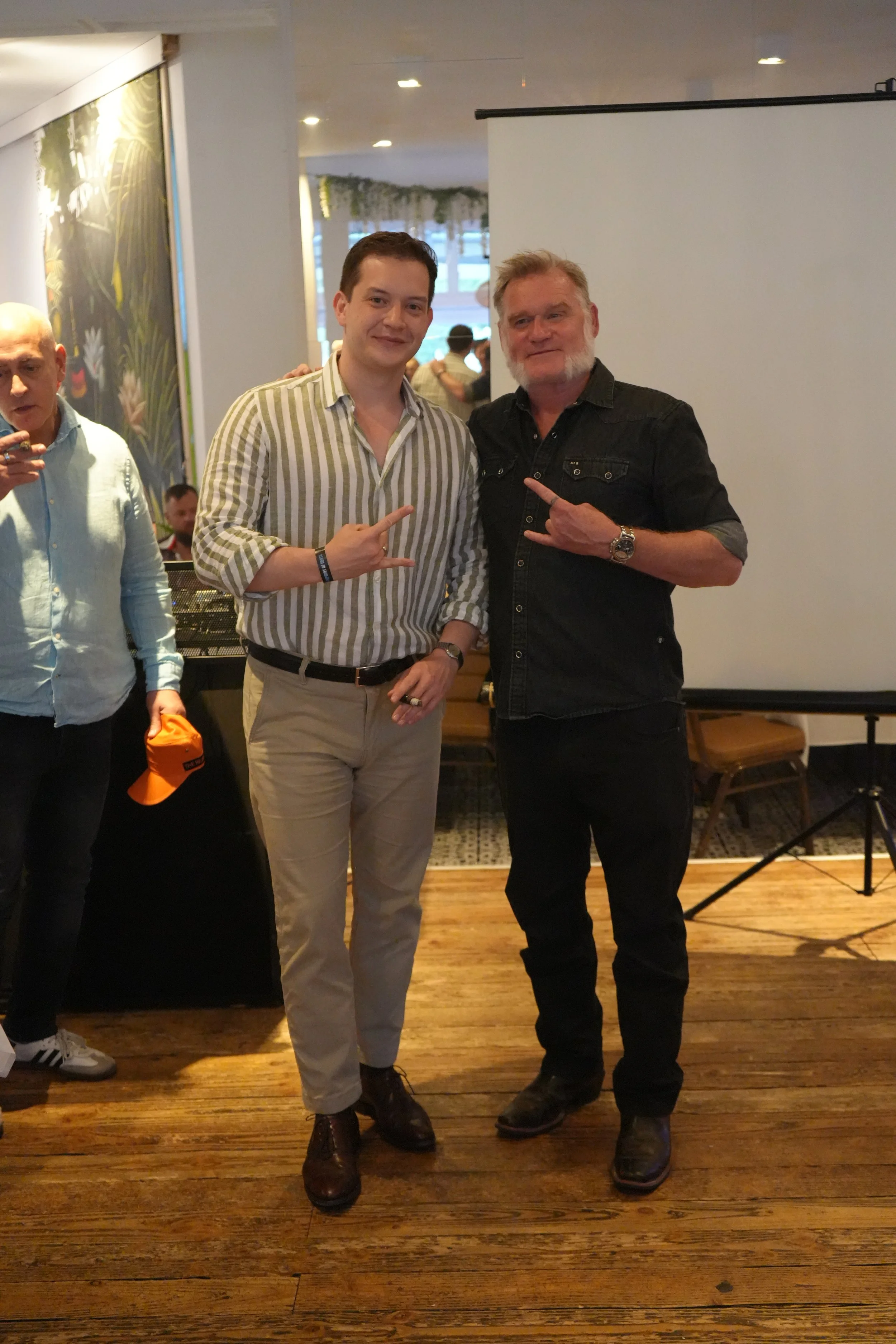 Two men standing next to each other, making hand gestures, with a third person partially visible on the left, in an indoor setting with wood flooring and a white screen.