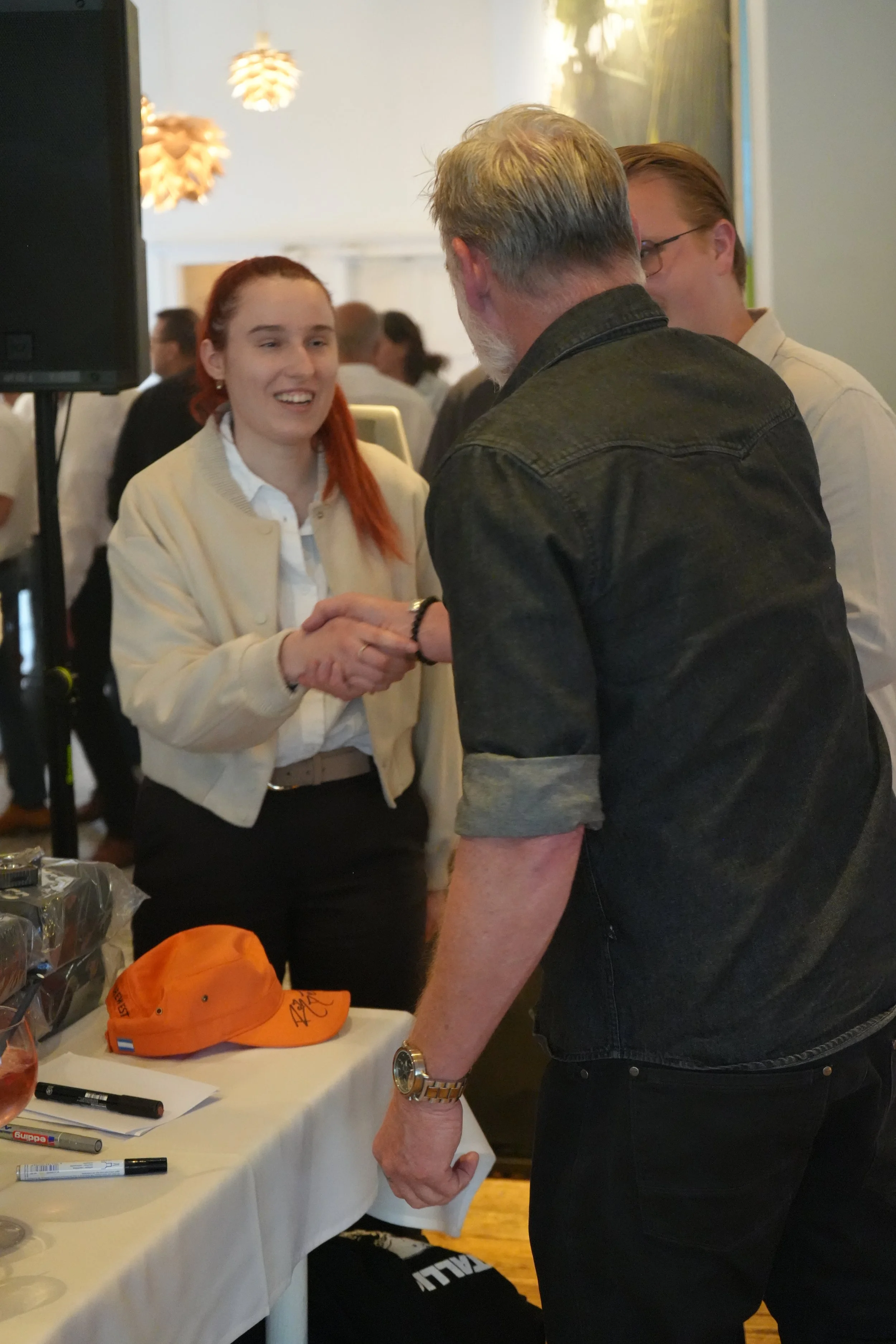 A young woman with red hair smiling and shaking hands with an older man with gray hair and a beard, wearing a black denim jacket at a social event or celebration.